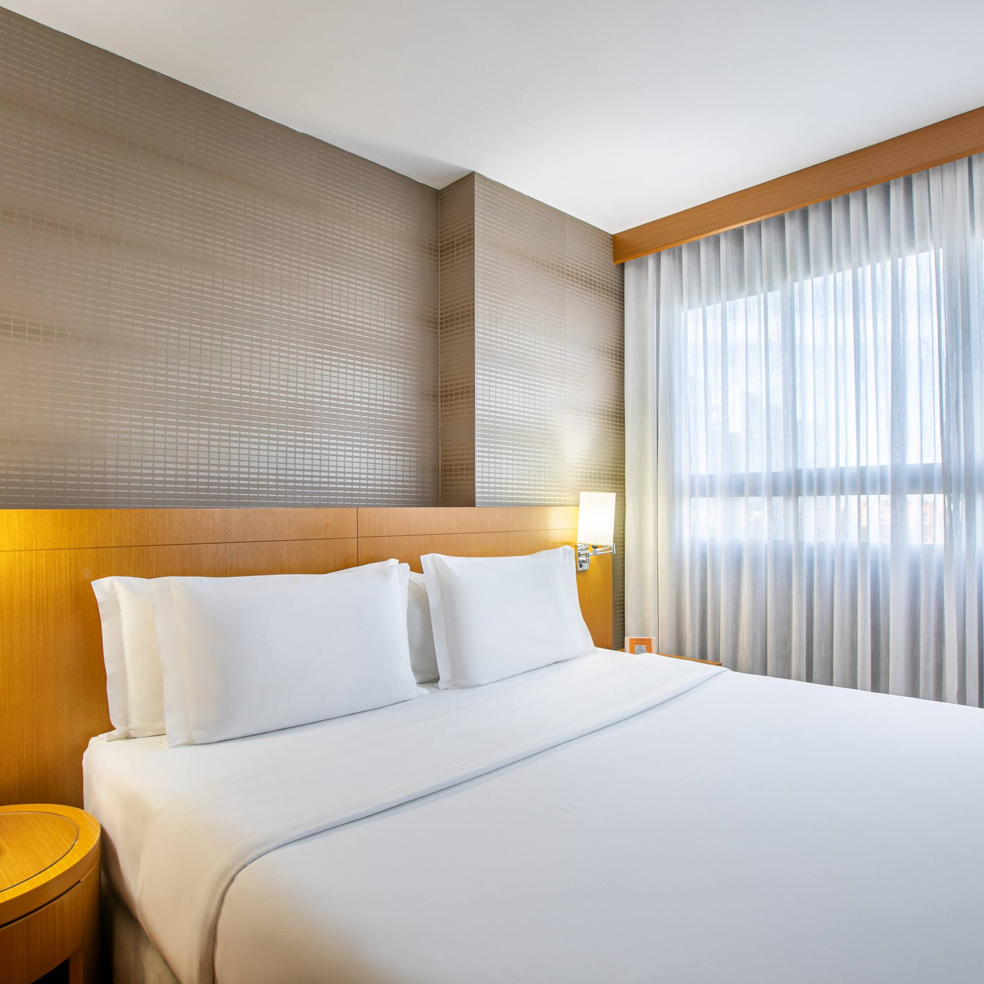 Modern hotel bedroom with white queen bed, wood headboard, and sheer curtains.