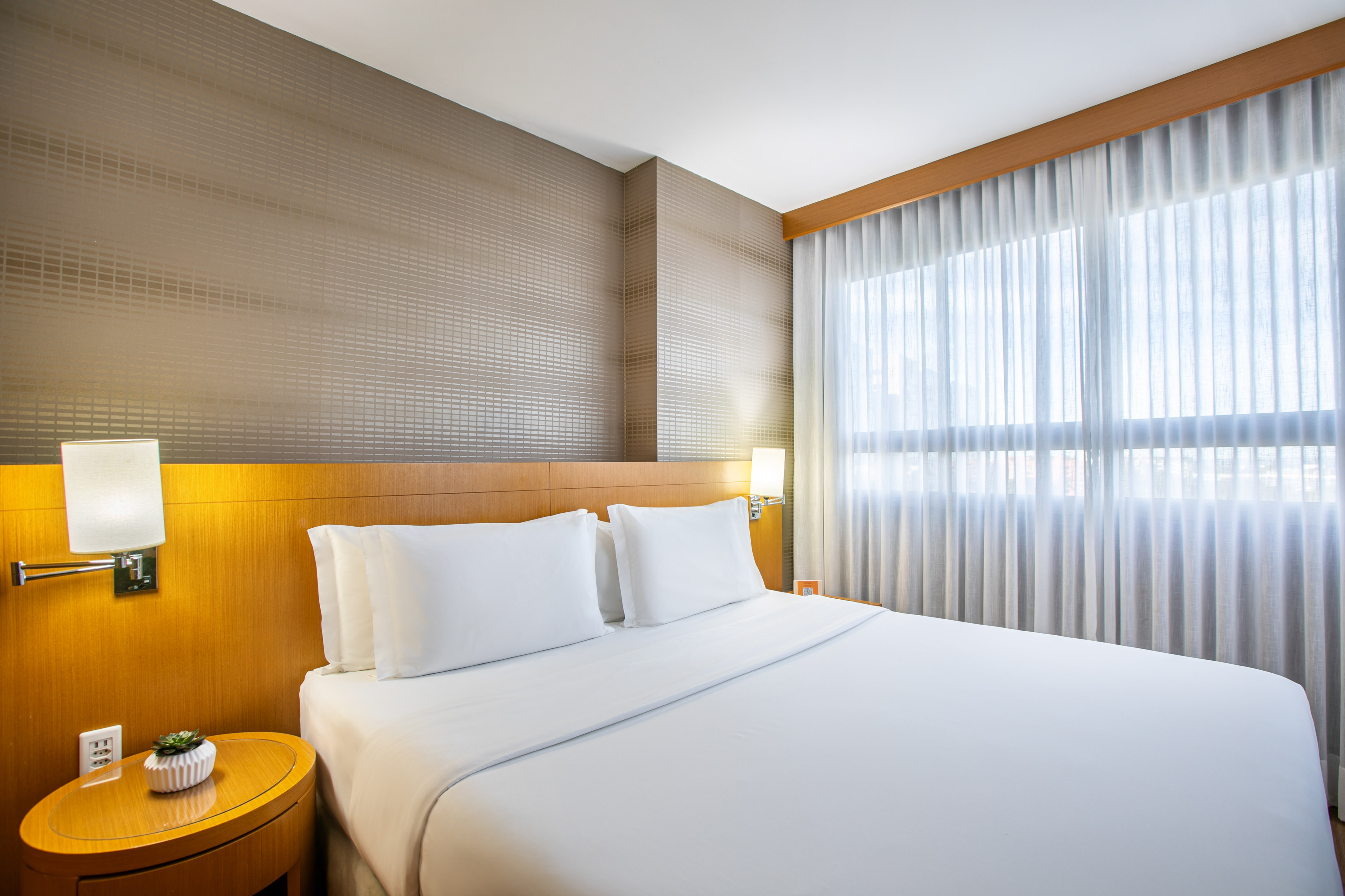 Modern hotel bedroom with white queen bed, wood headboard, and sheer curtains.