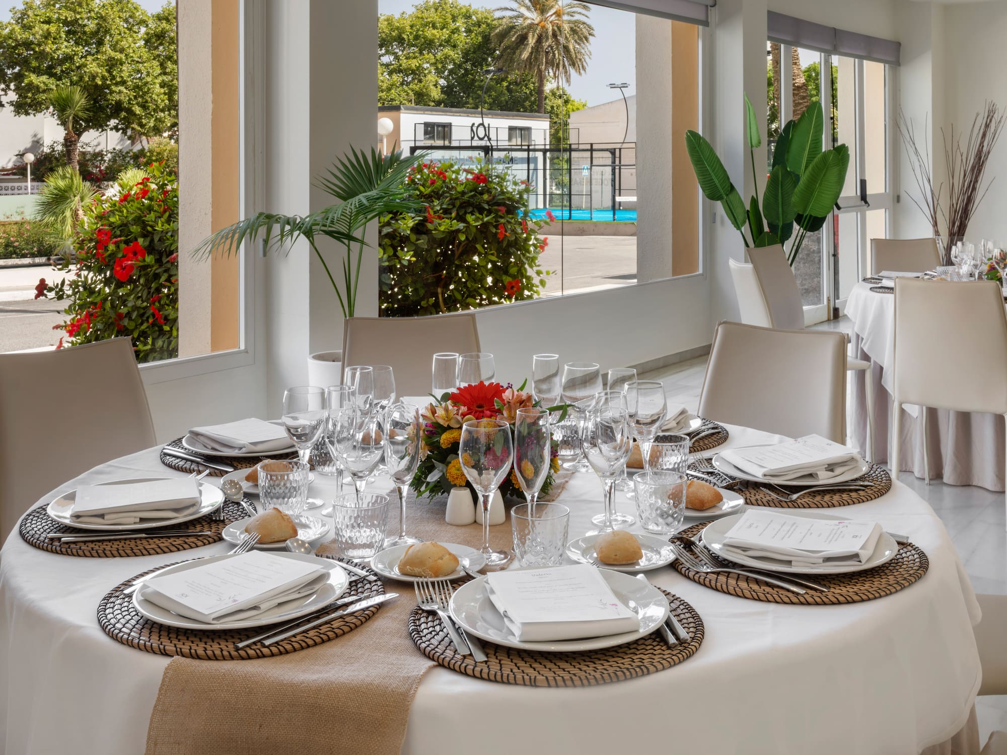Mesa de restaurante preparada con centro floral y vista a cancha de pádel.