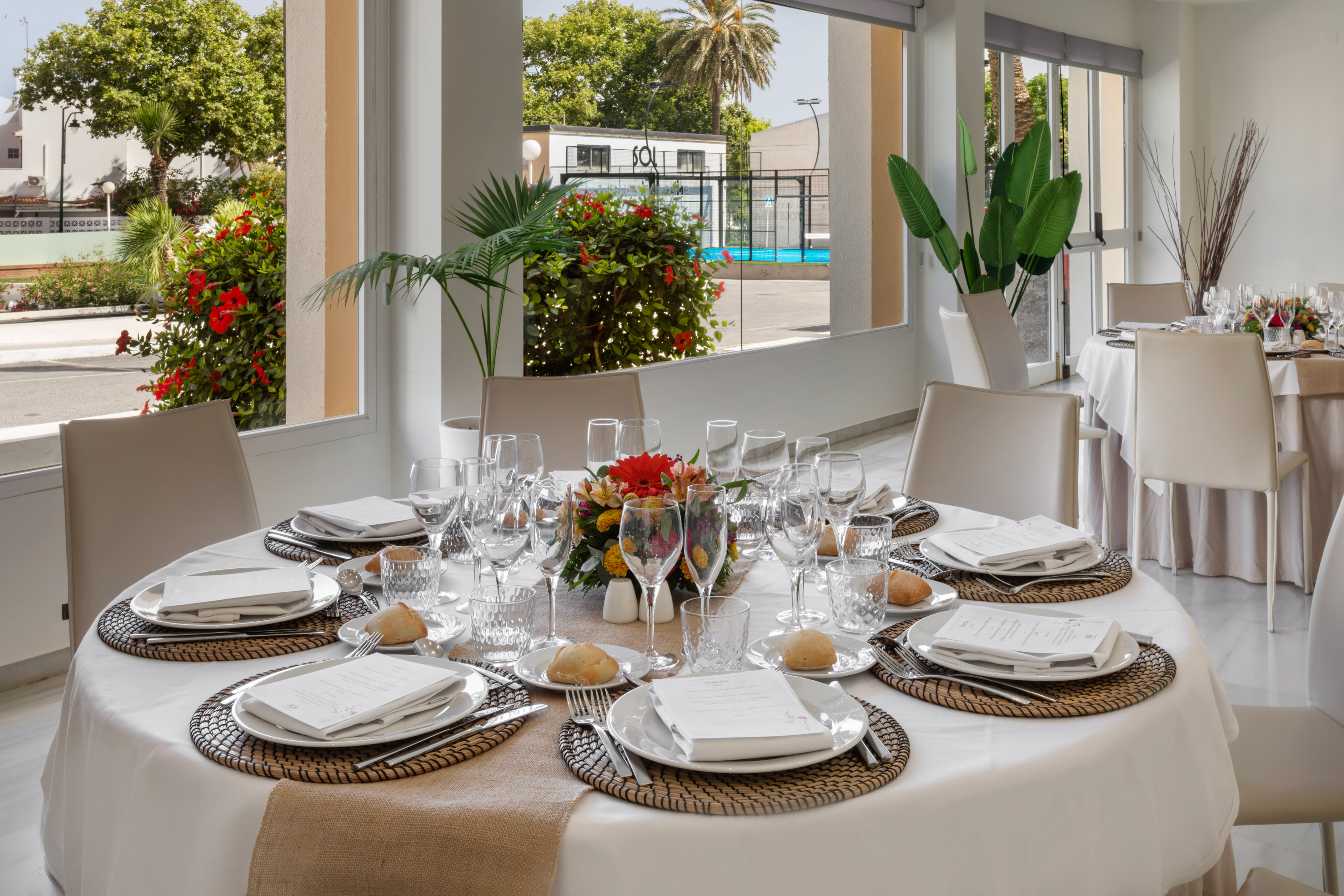 Mesa de restaurante preparada con centro floral y vista a cancha de pádel.
