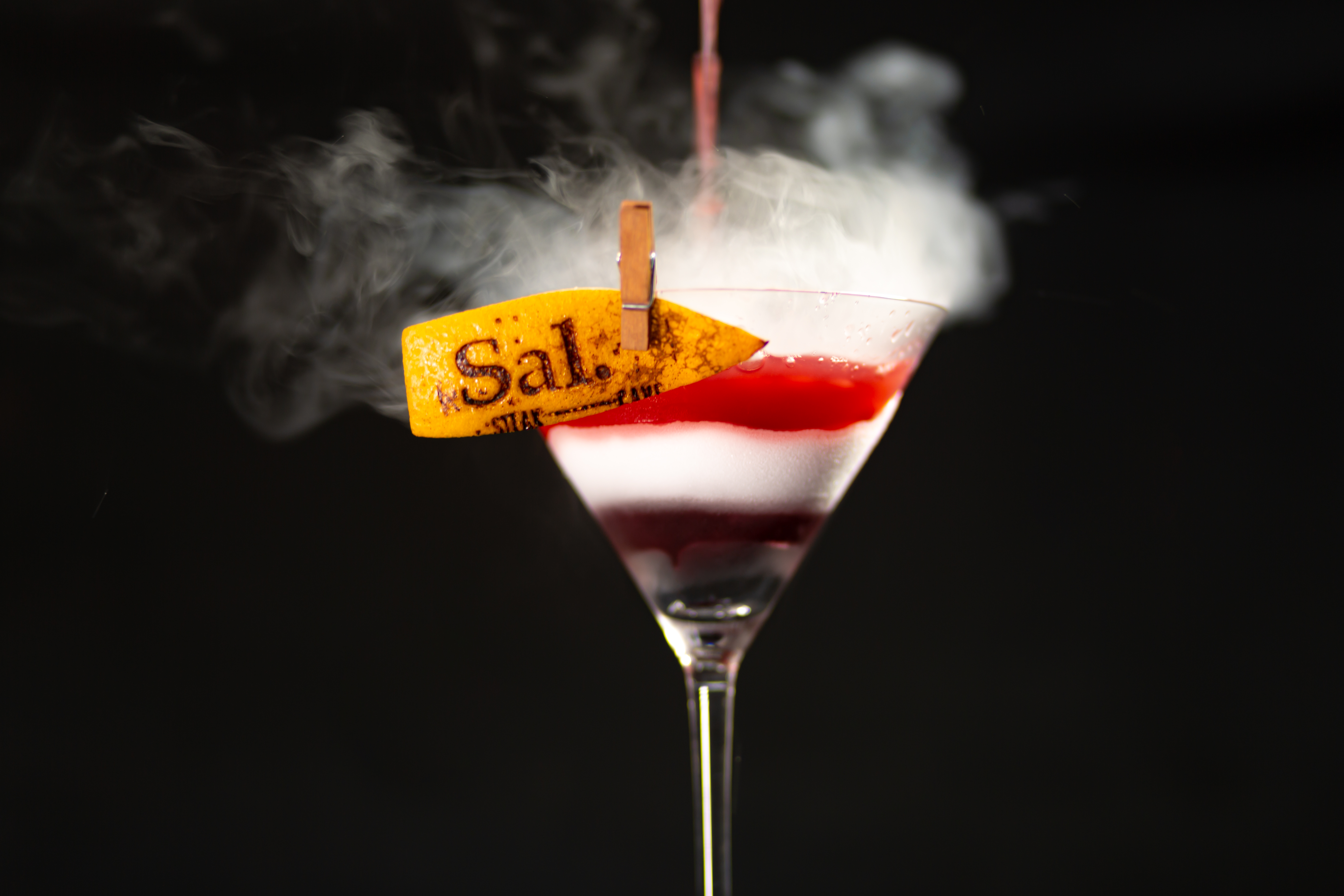 Smoky layered martini cocktail, red & white, with 'Sal.' orange peel garnish.