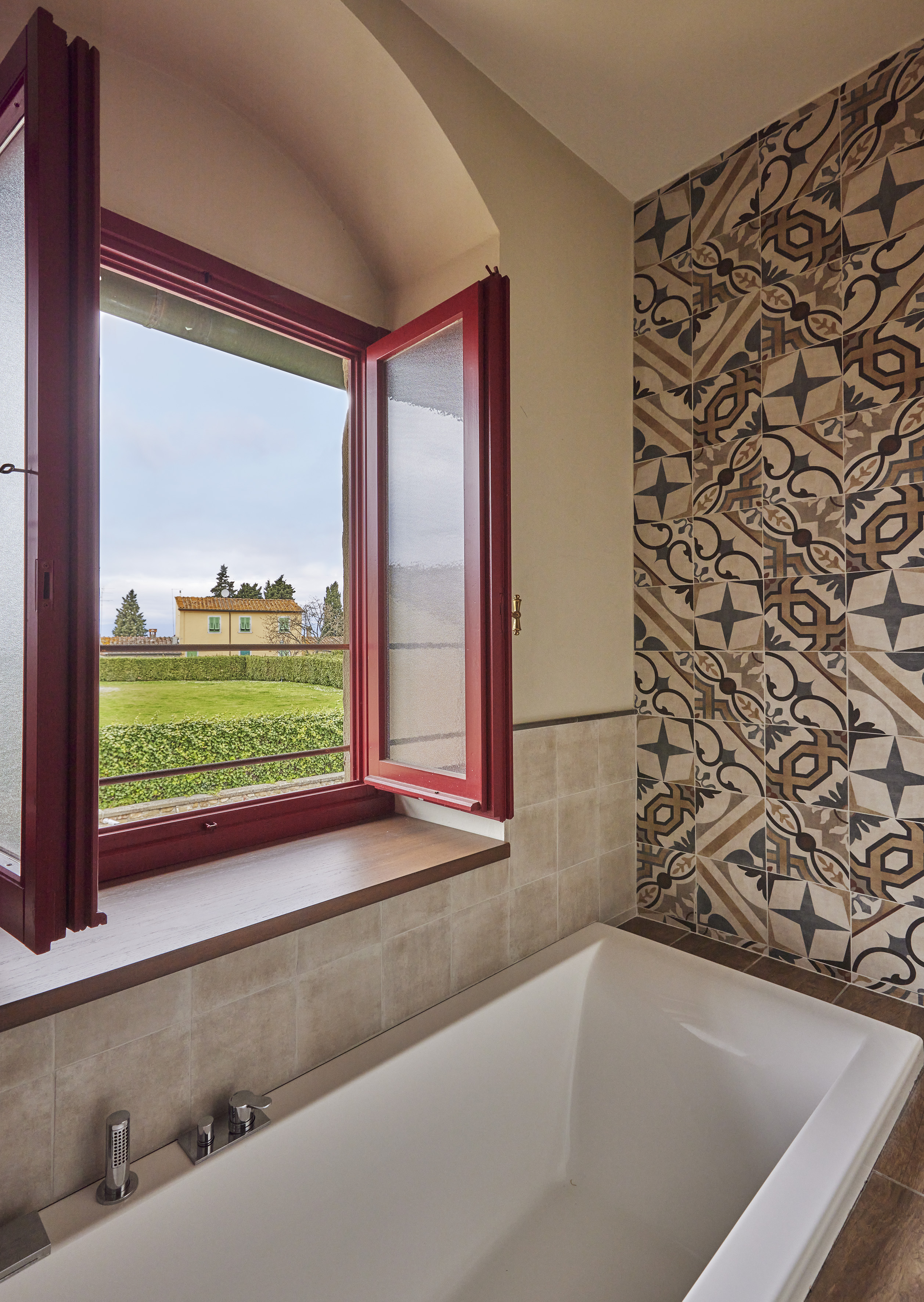 Bathtub and patterned bathroom tiles with a red window overlooking a green garden view.