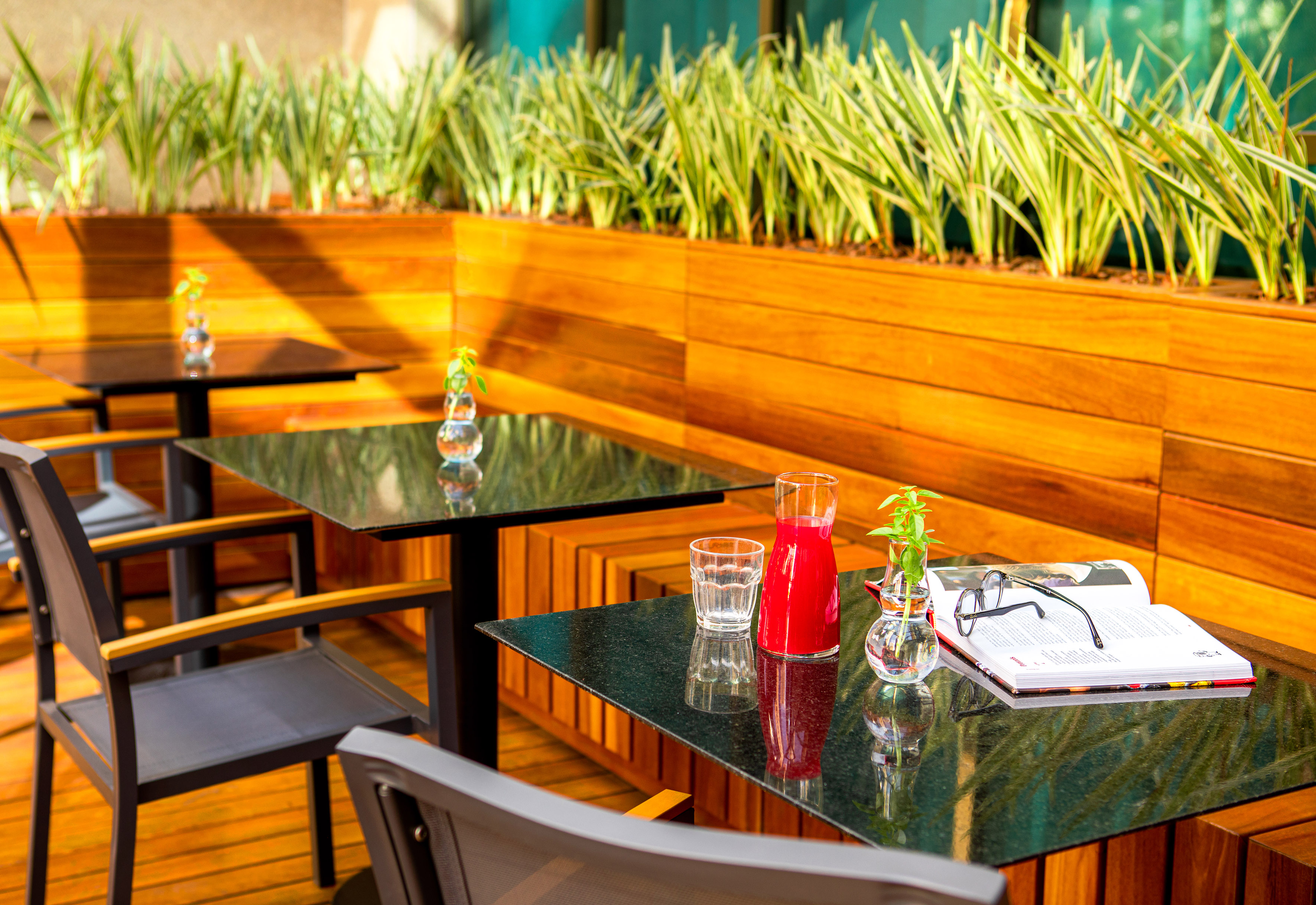 Outdoor cafe with dark tables, red juice, book, glasses, and lush green plants.