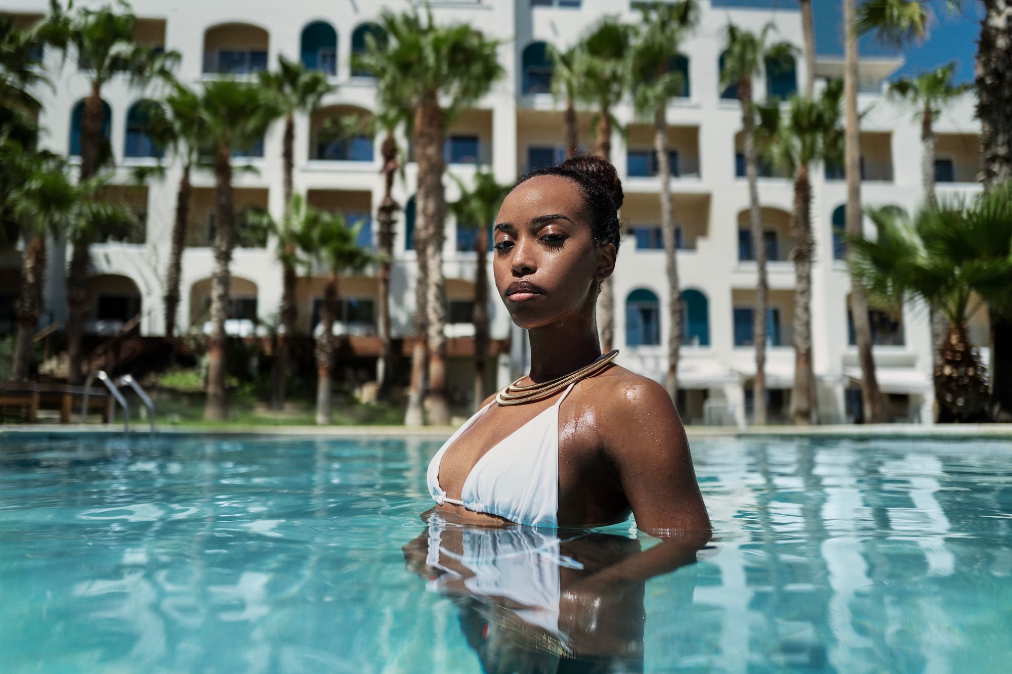 Woman in white bikini, gold necklace, swimming in resort pool with palm trees.