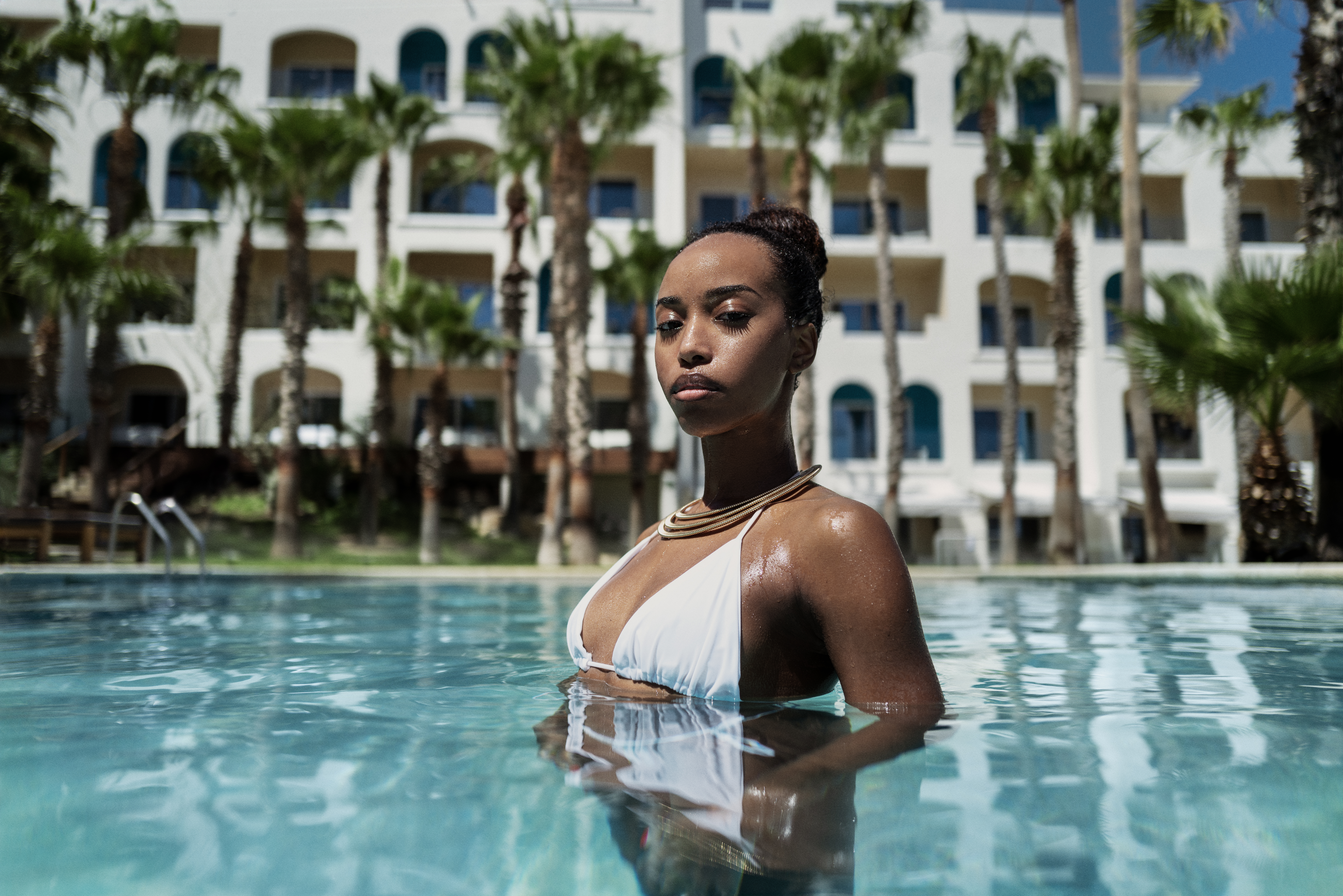 Woman in white bikini, gold necklace, swimming in resort pool with palm trees.