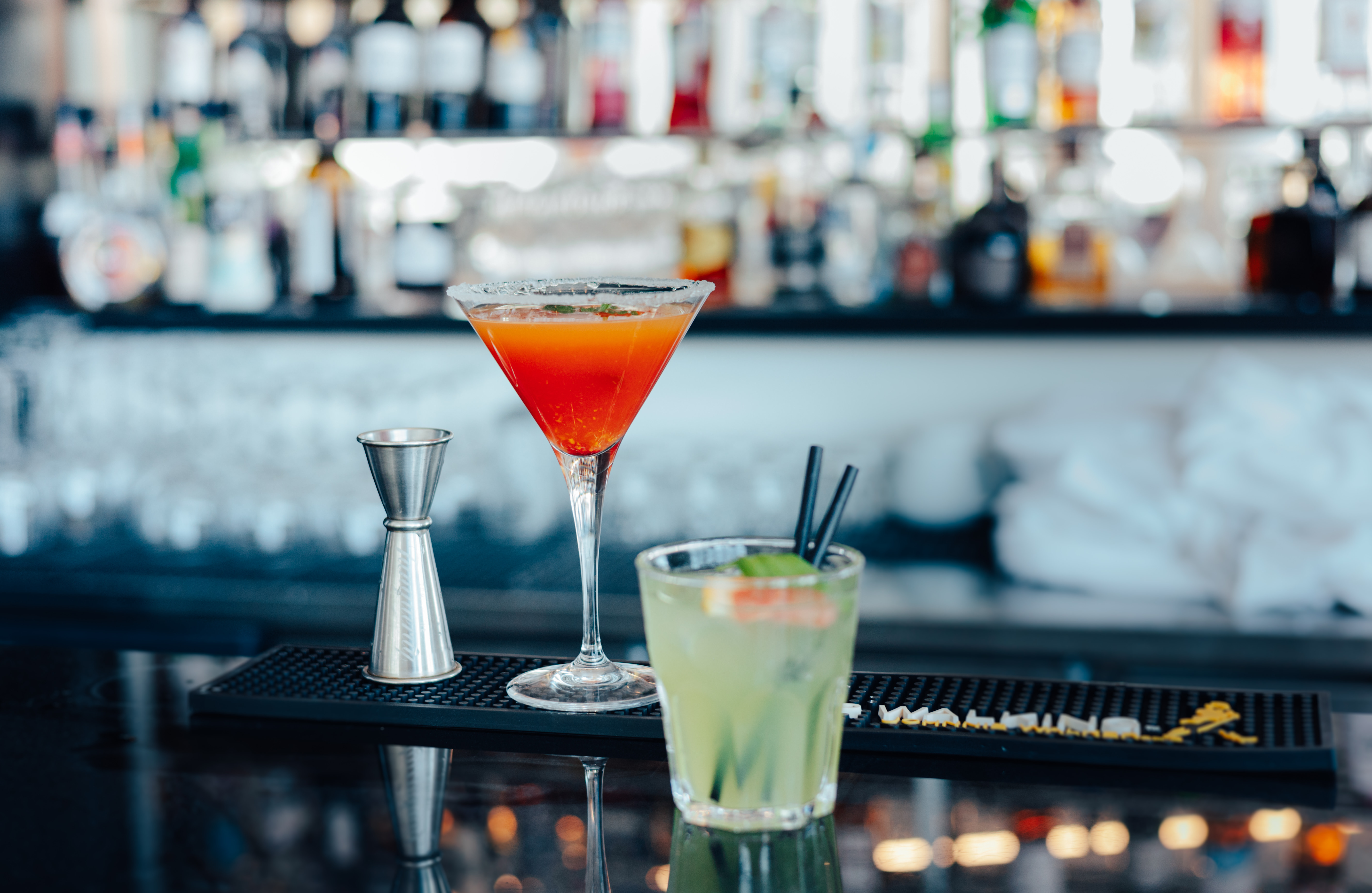 Two cocktails: a red martini and green drink with a jigger on a bar.