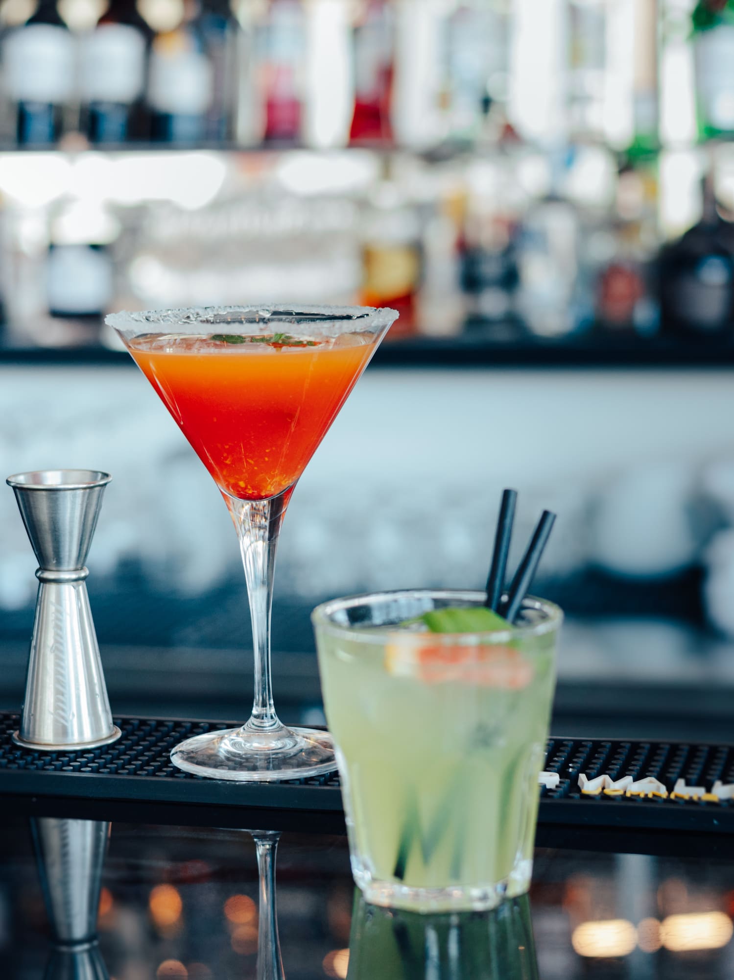 Two cocktails: a red martini and green drink with a jigger on a bar.