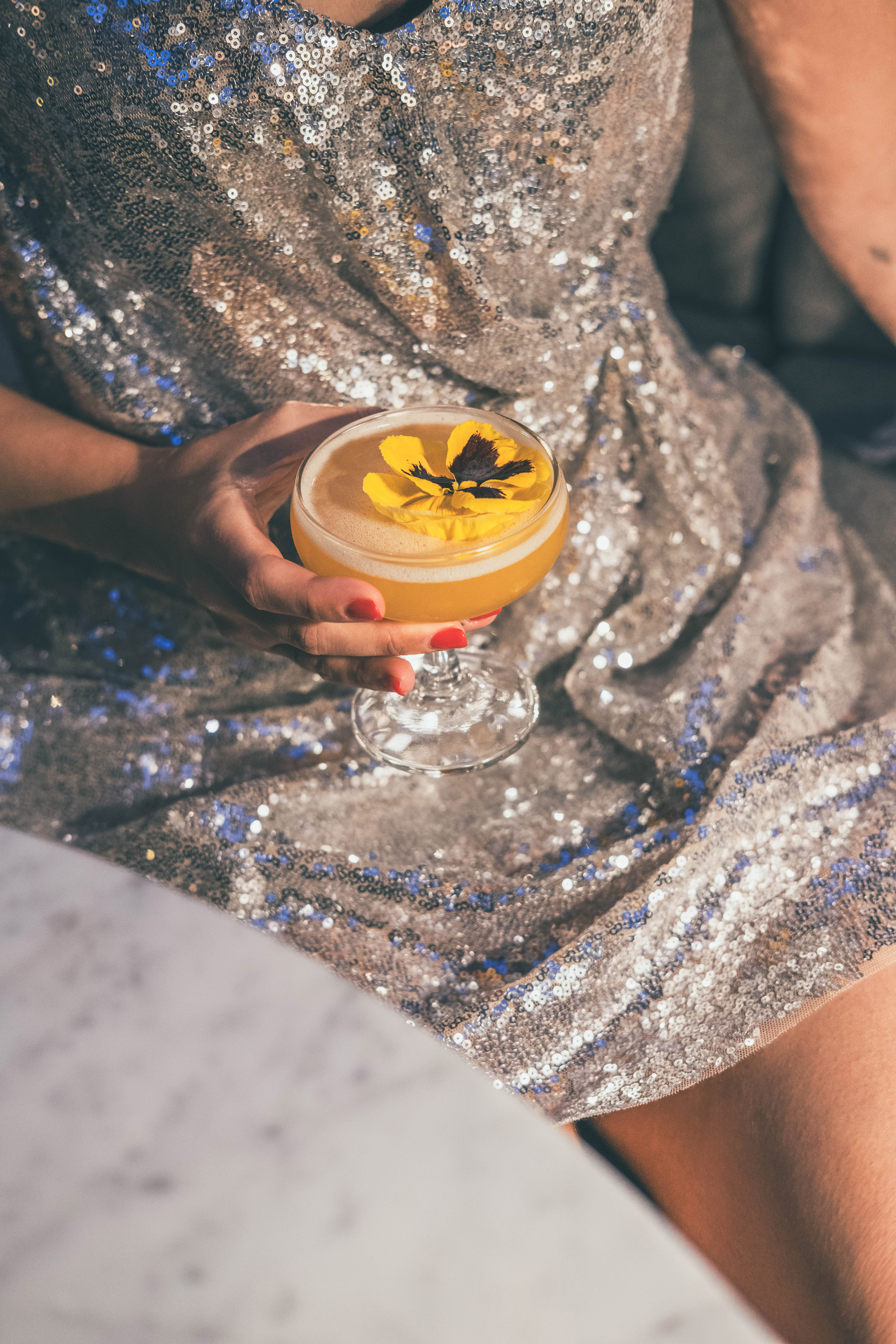 Hand in sequin dress holding yellow cocktail with a pansy garnish.