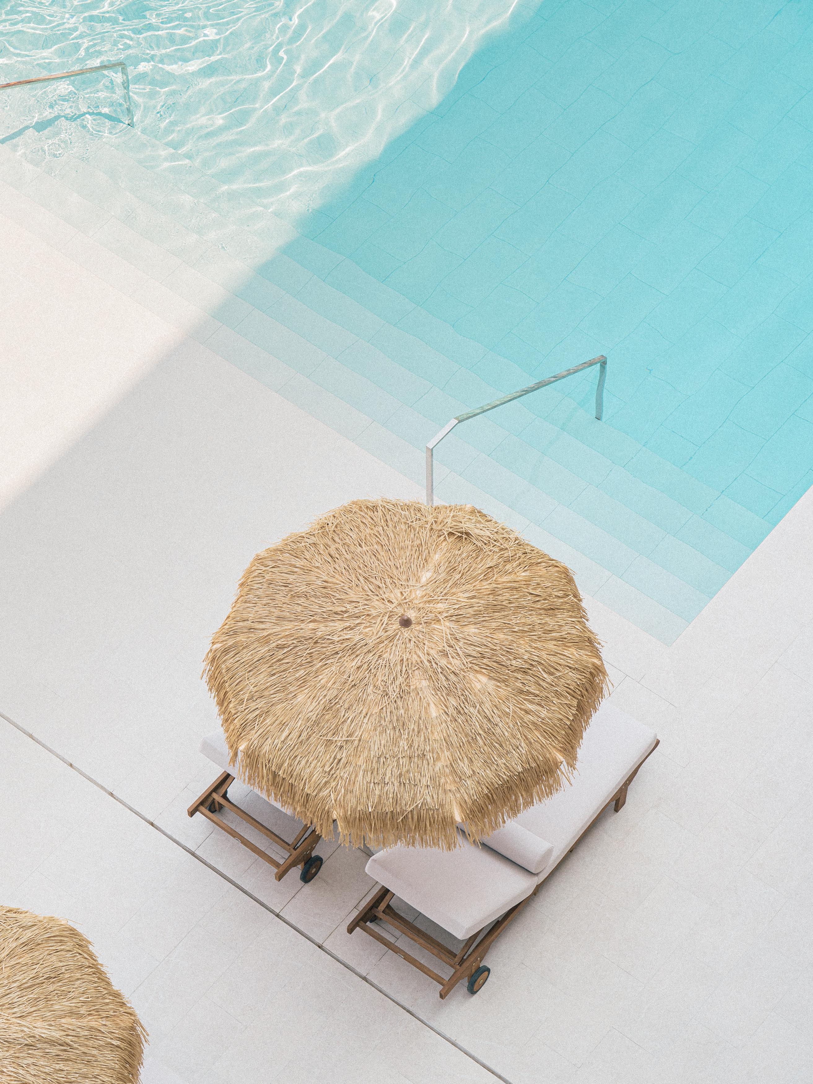 Piscina azul con escalones, sombrilla de paja y tumbonas.