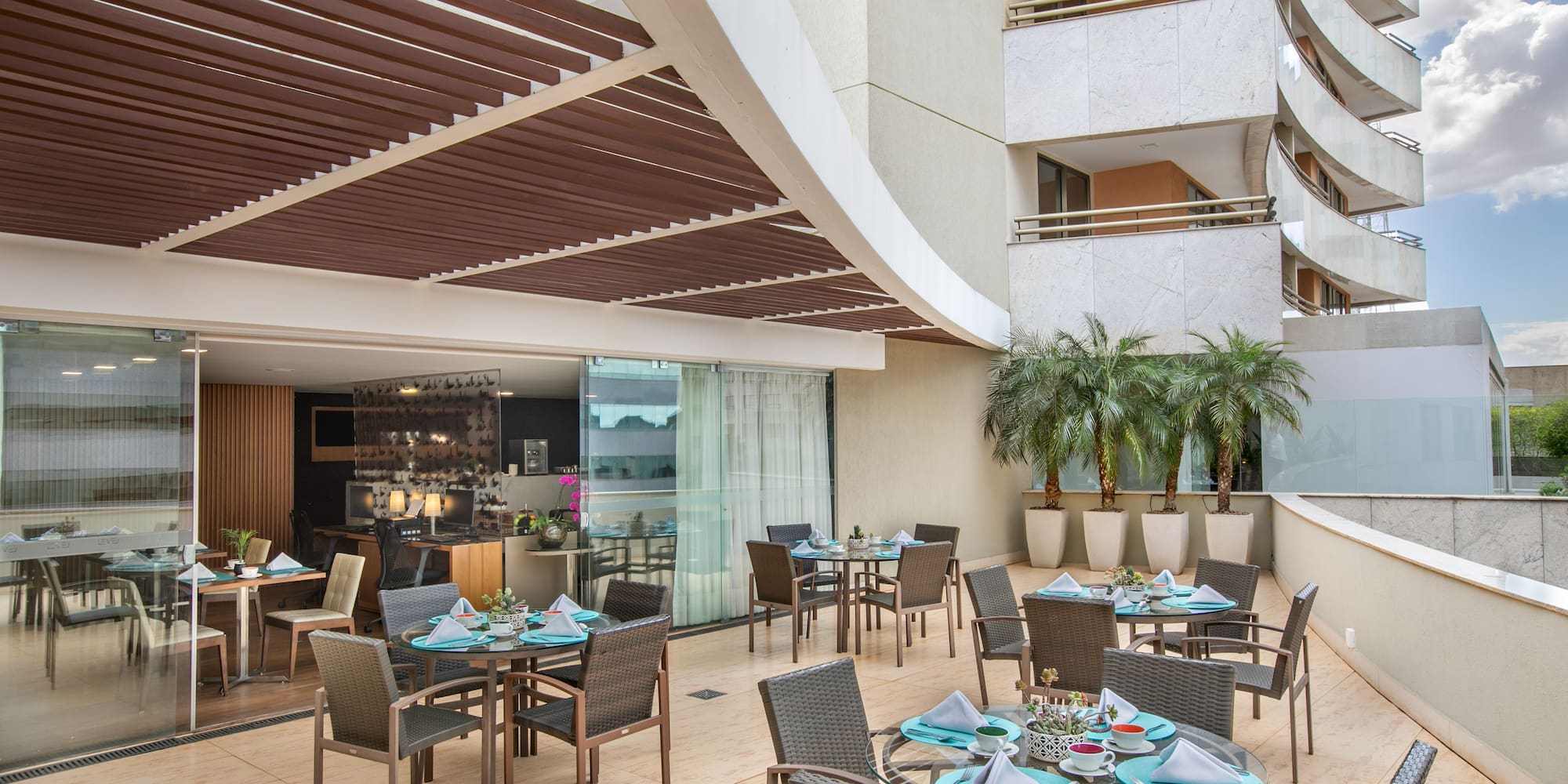 Modern outdoor dining terrace with set tables, chairs, and palm trees.
