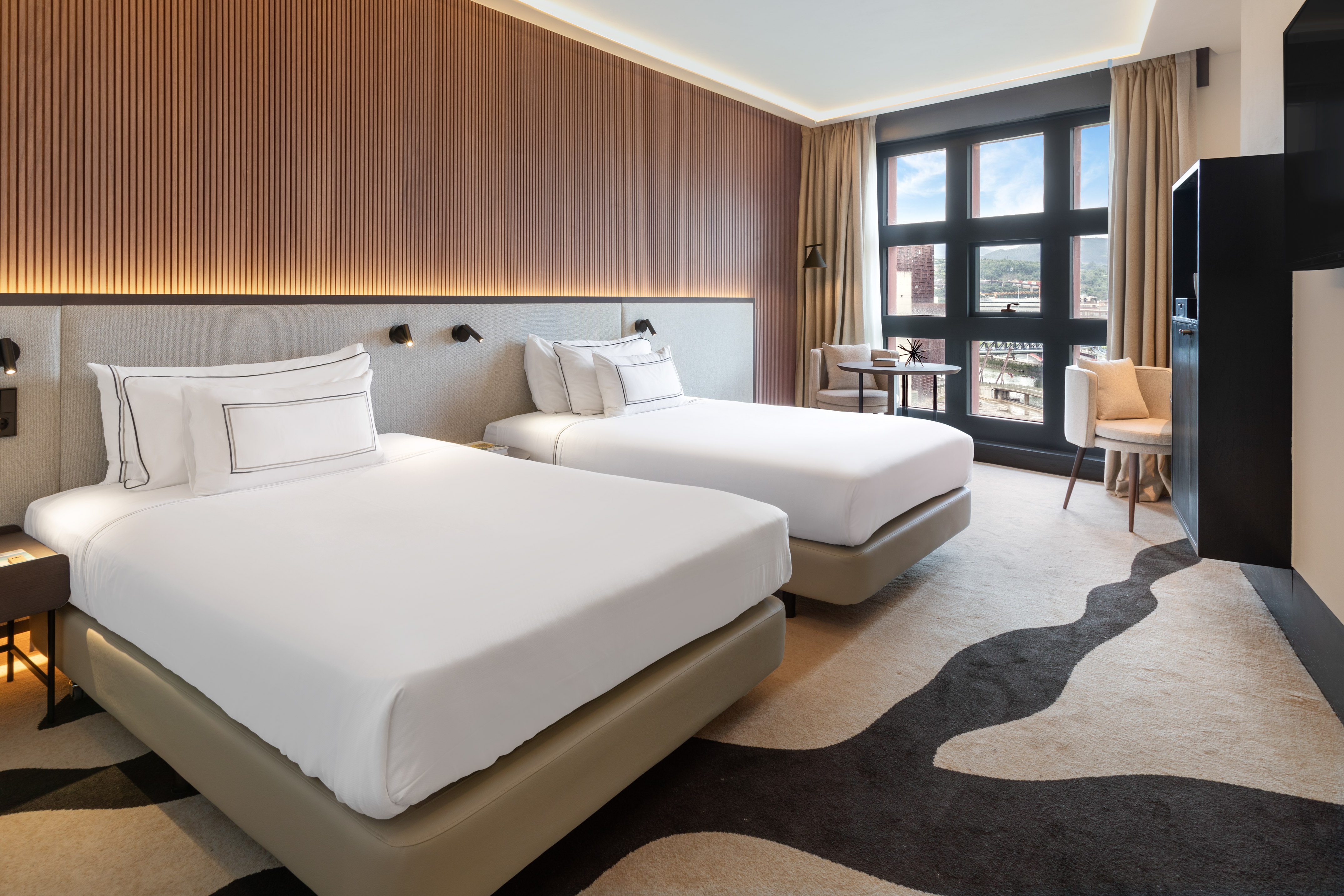 Modern hotel twin room with wood wall, two beds, window view, patterned carpet.