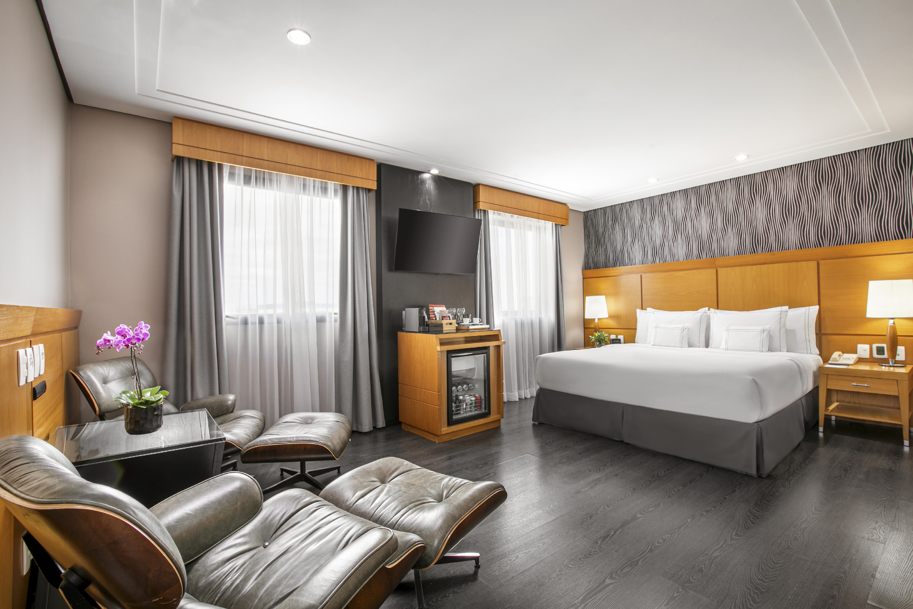 Modern hotel bedroom with king bed, lounge chairs, TV, and minibar.