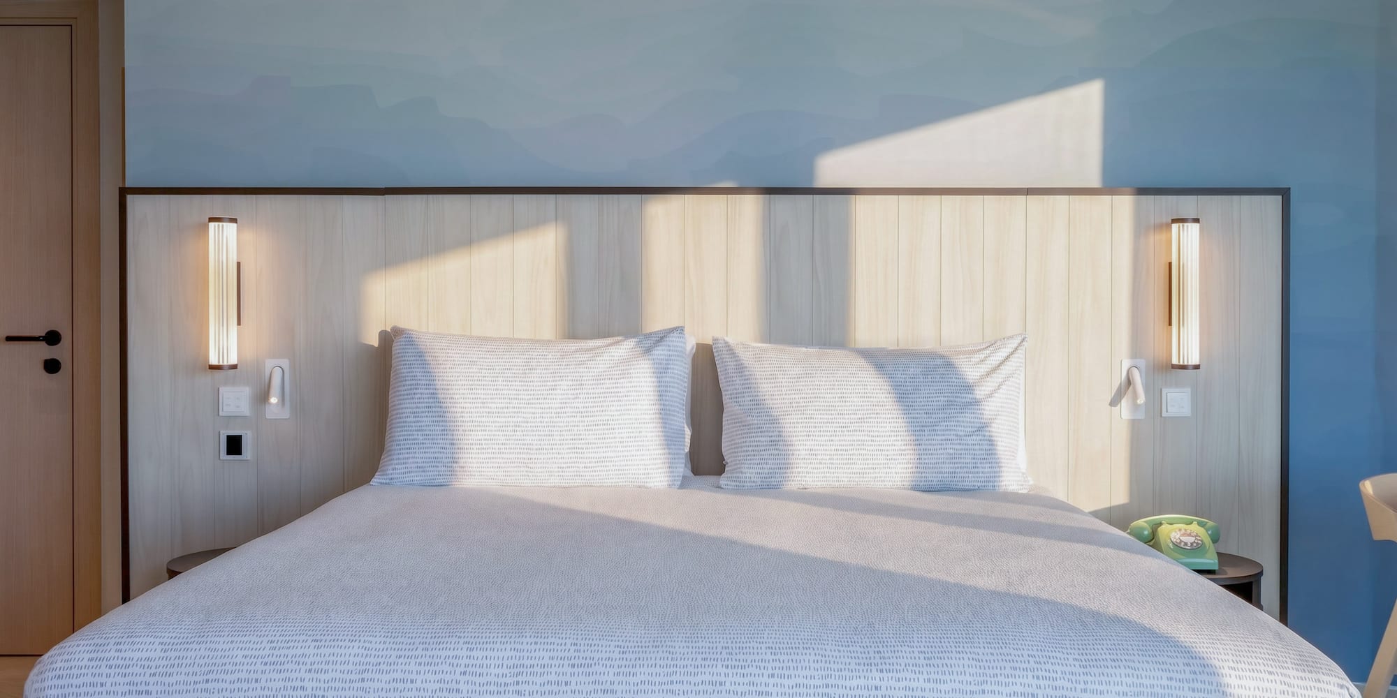 Modern hotel bedroom with sunlit patterned bed, wooden headboard, and blue wall.