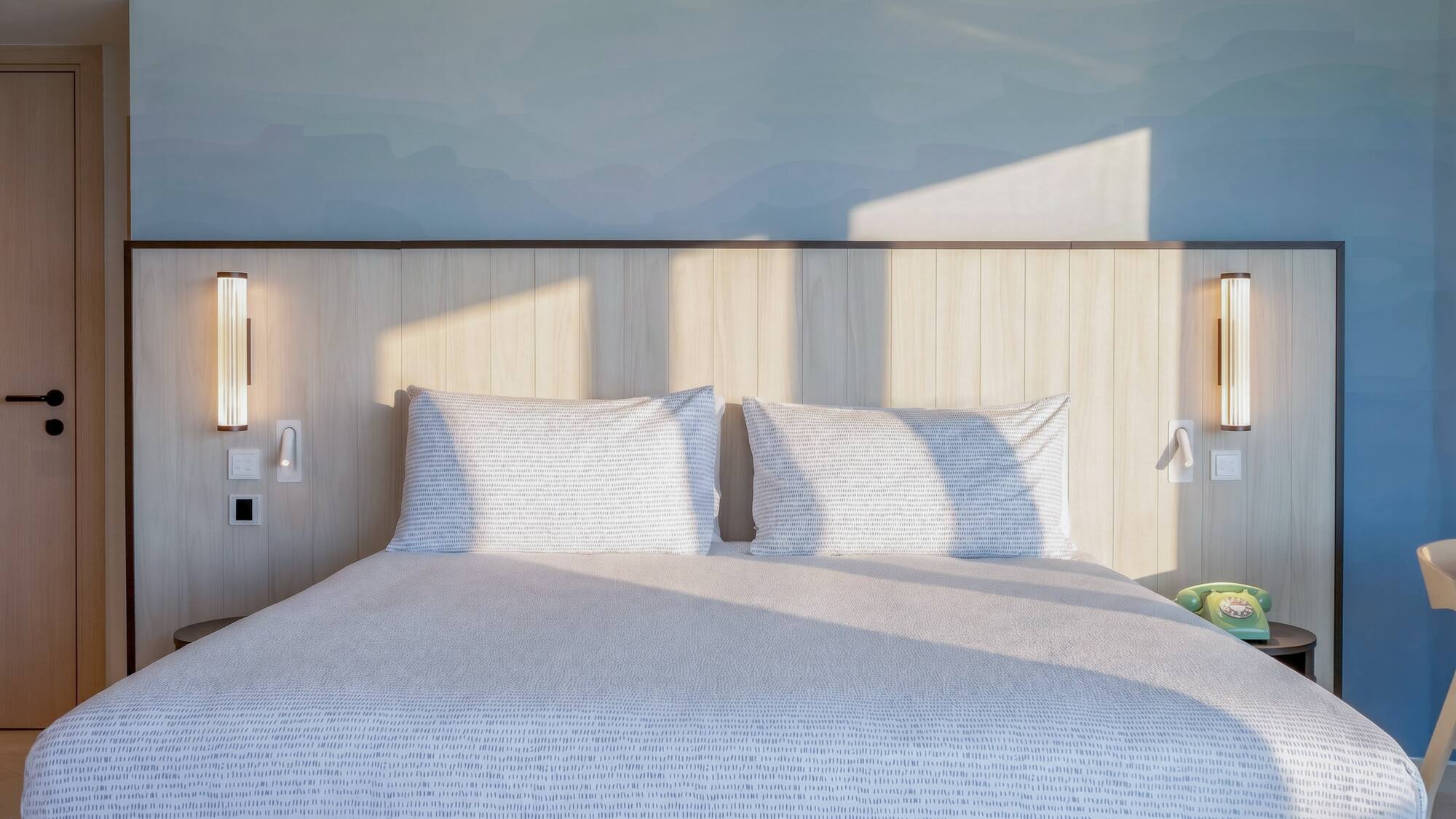 Modern hotel bedroom with sunlit patterned bed, wooden headboard, and blue wall.