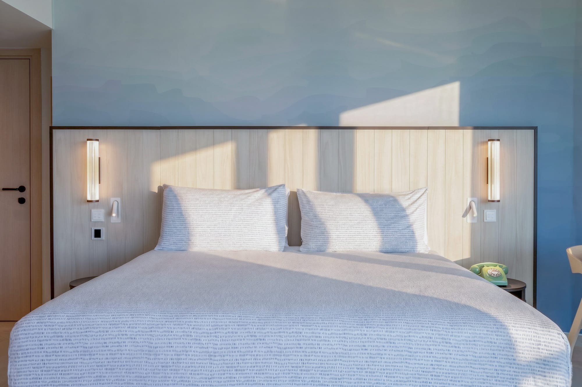 Modern hotel bedroom with sunlit patterned bed, wooden headboard, and blue wall.