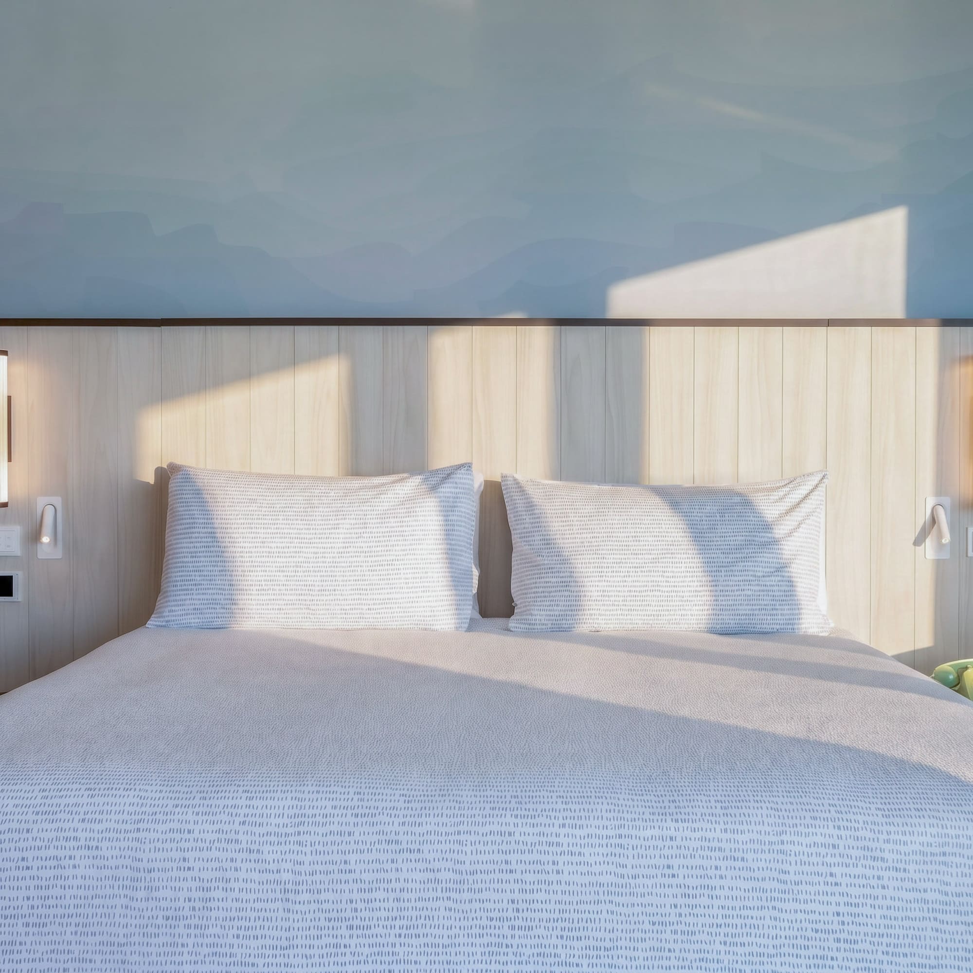 Modern hotel bedroom with sunlit patterned bed, wooden headboard, and blue wall.