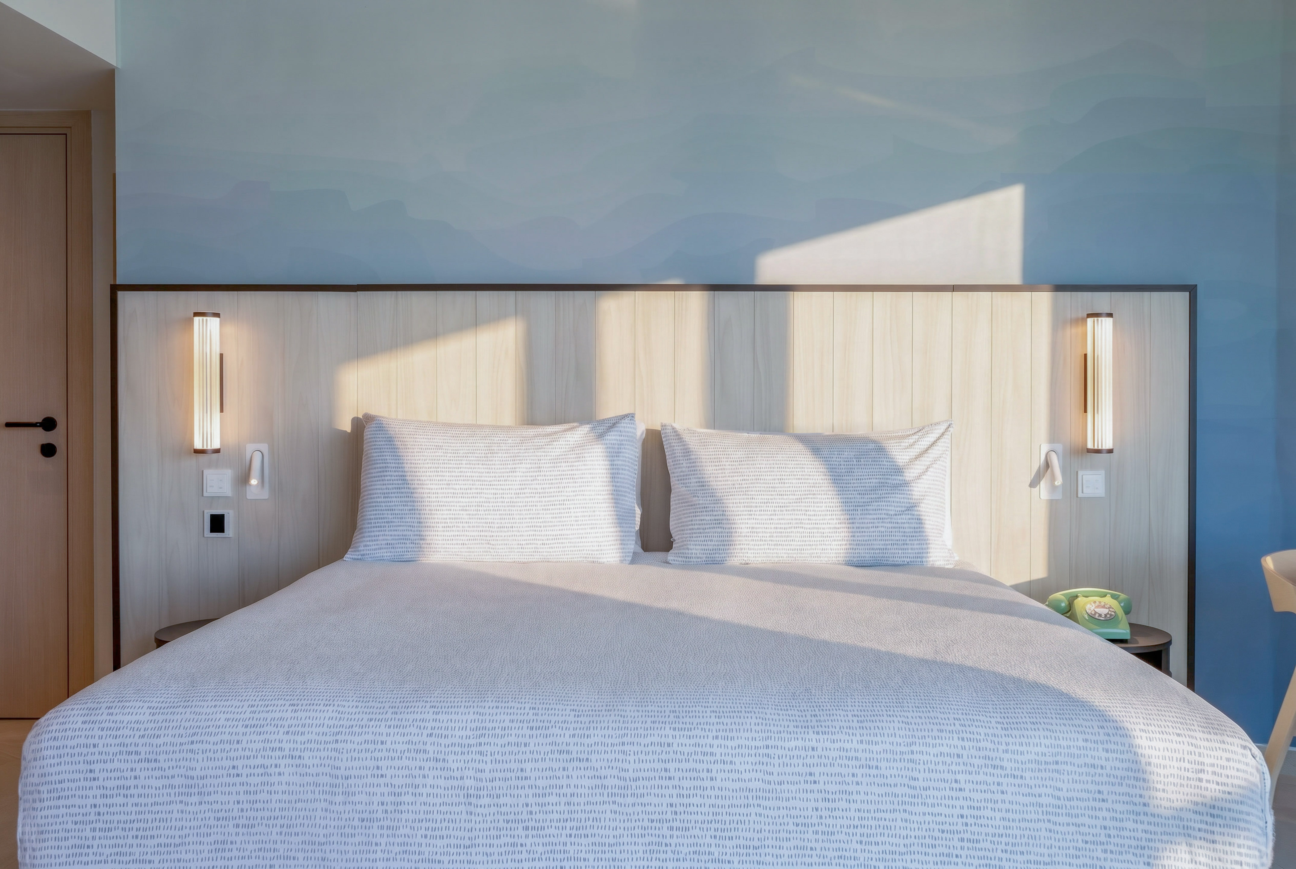 Modern hotel bedroom with sunlit patterned bed, wooden headboard, and blue wall.
