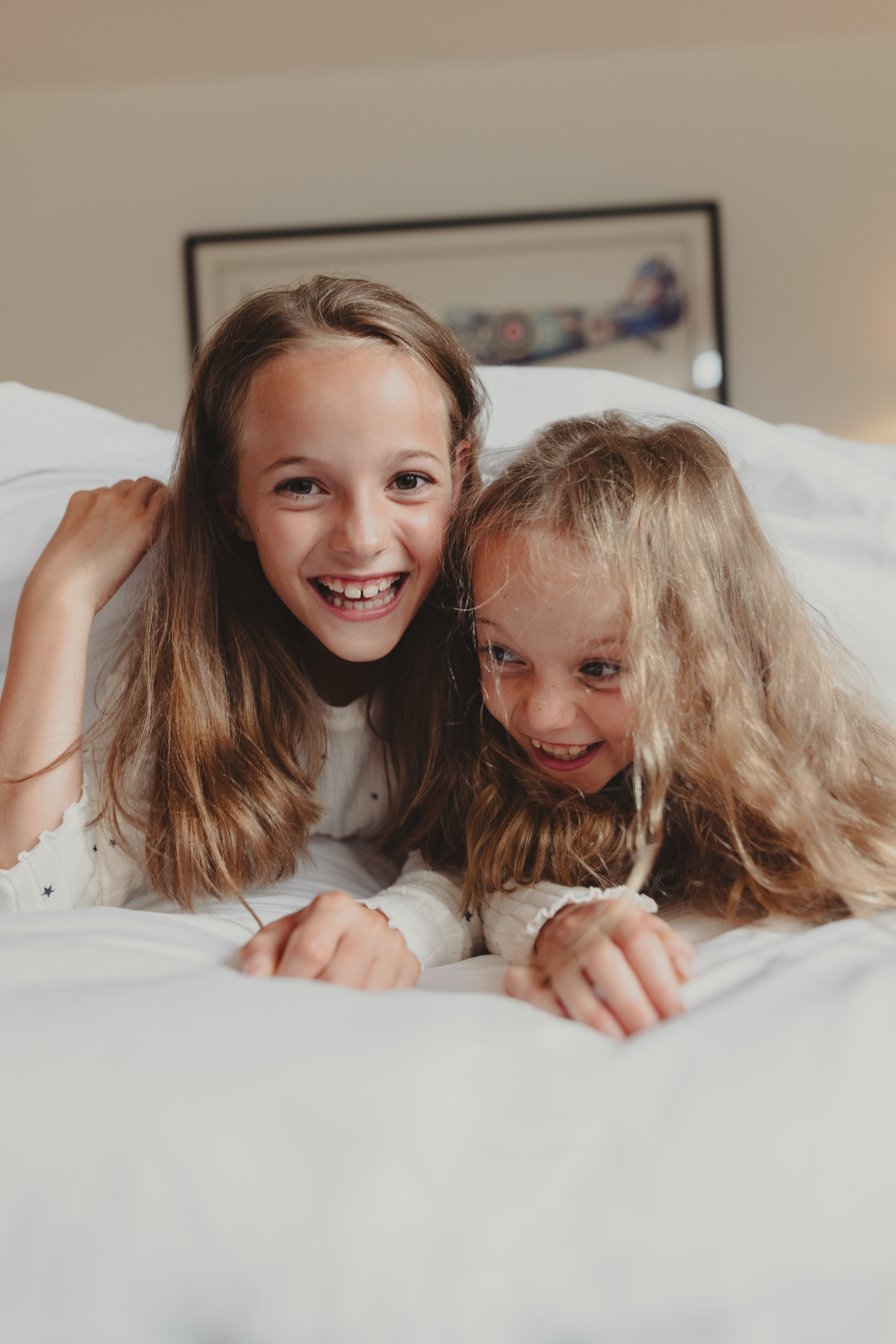 Two happy girls laughing playfully on a white bed, morning.