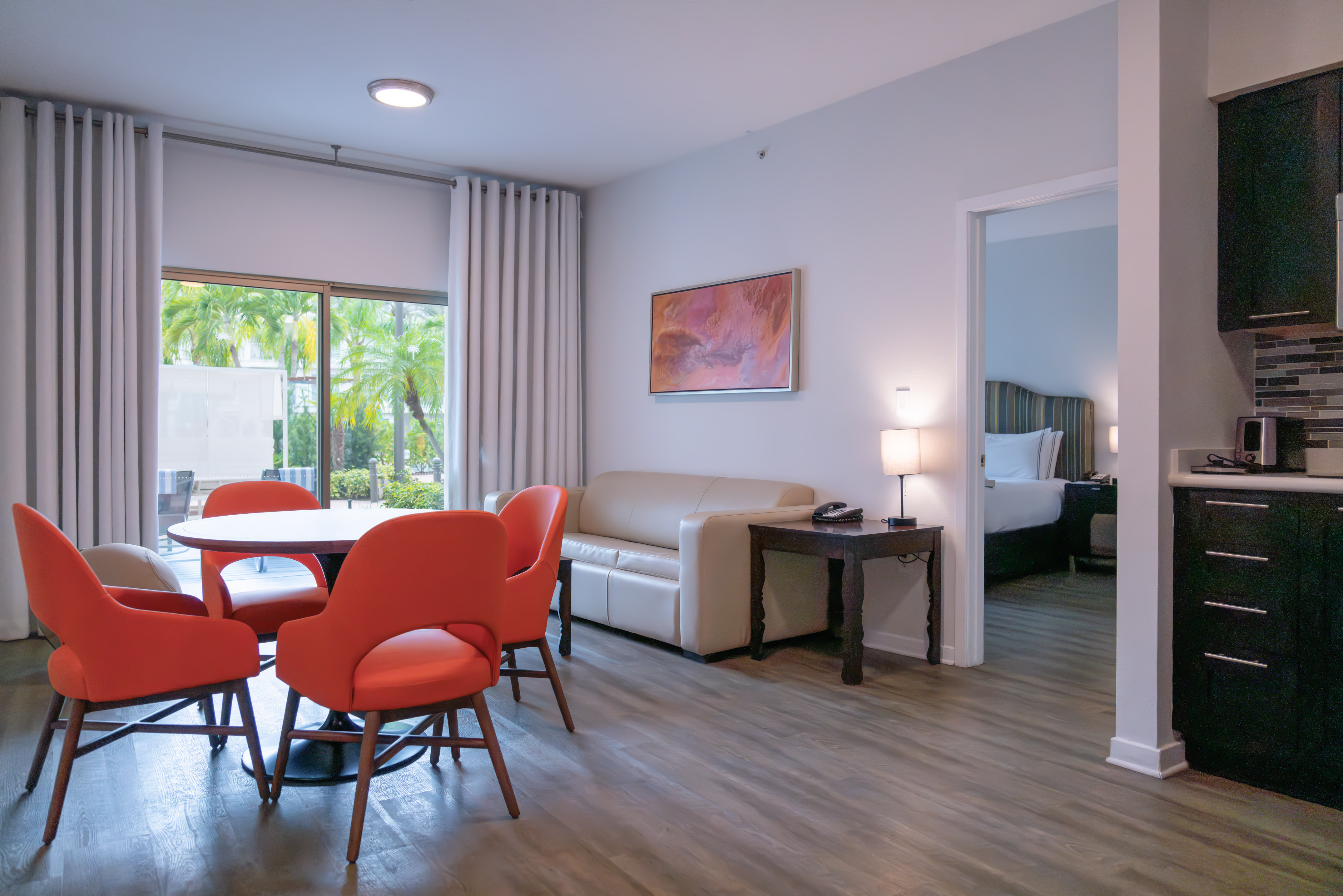 Modern hotel suite living area with dining table, orange chairs, sofa, and bedroom.