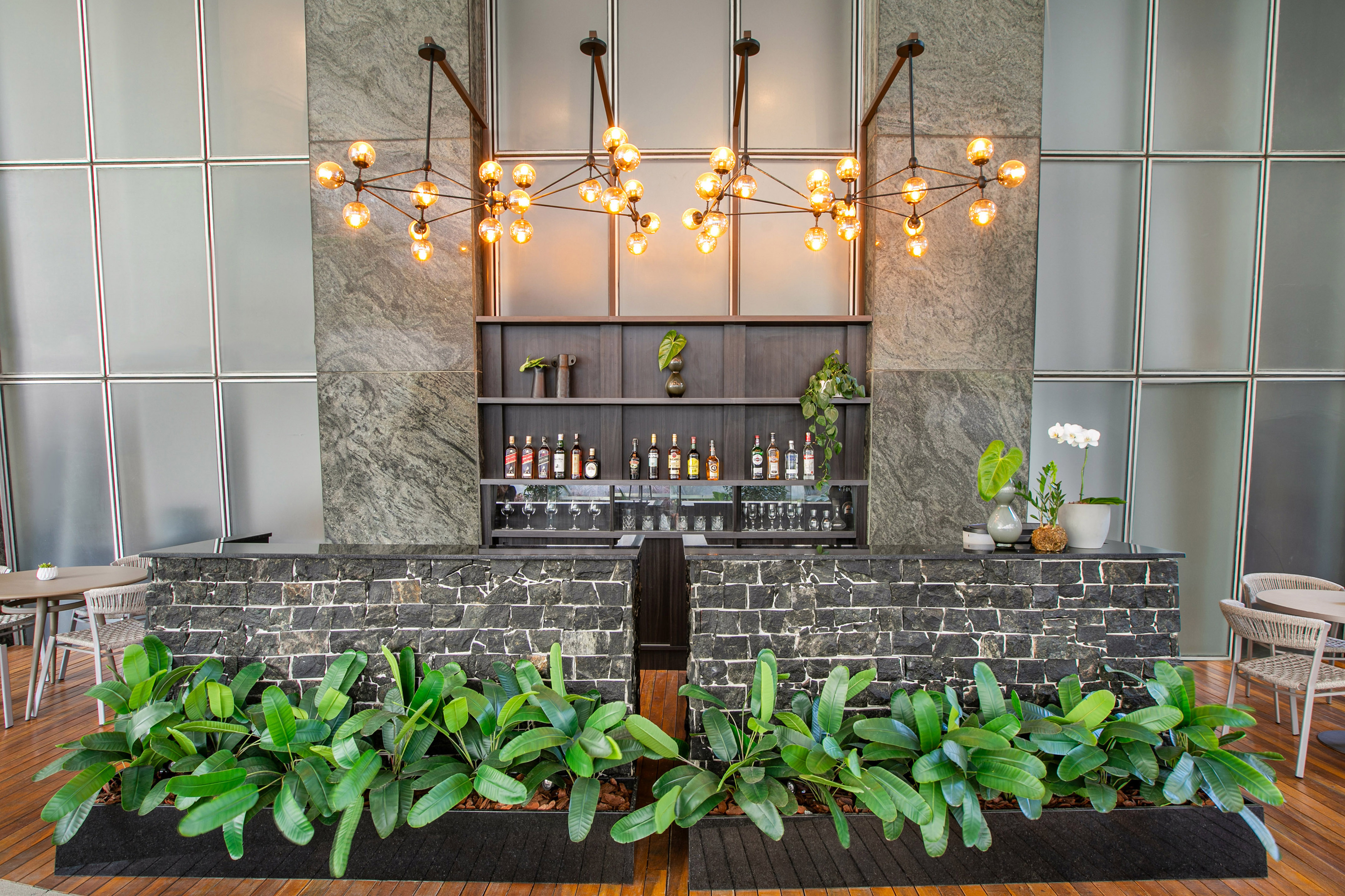 Modern hotel bar, stone counter, liquor bottles, bright chandeliers, green plants.