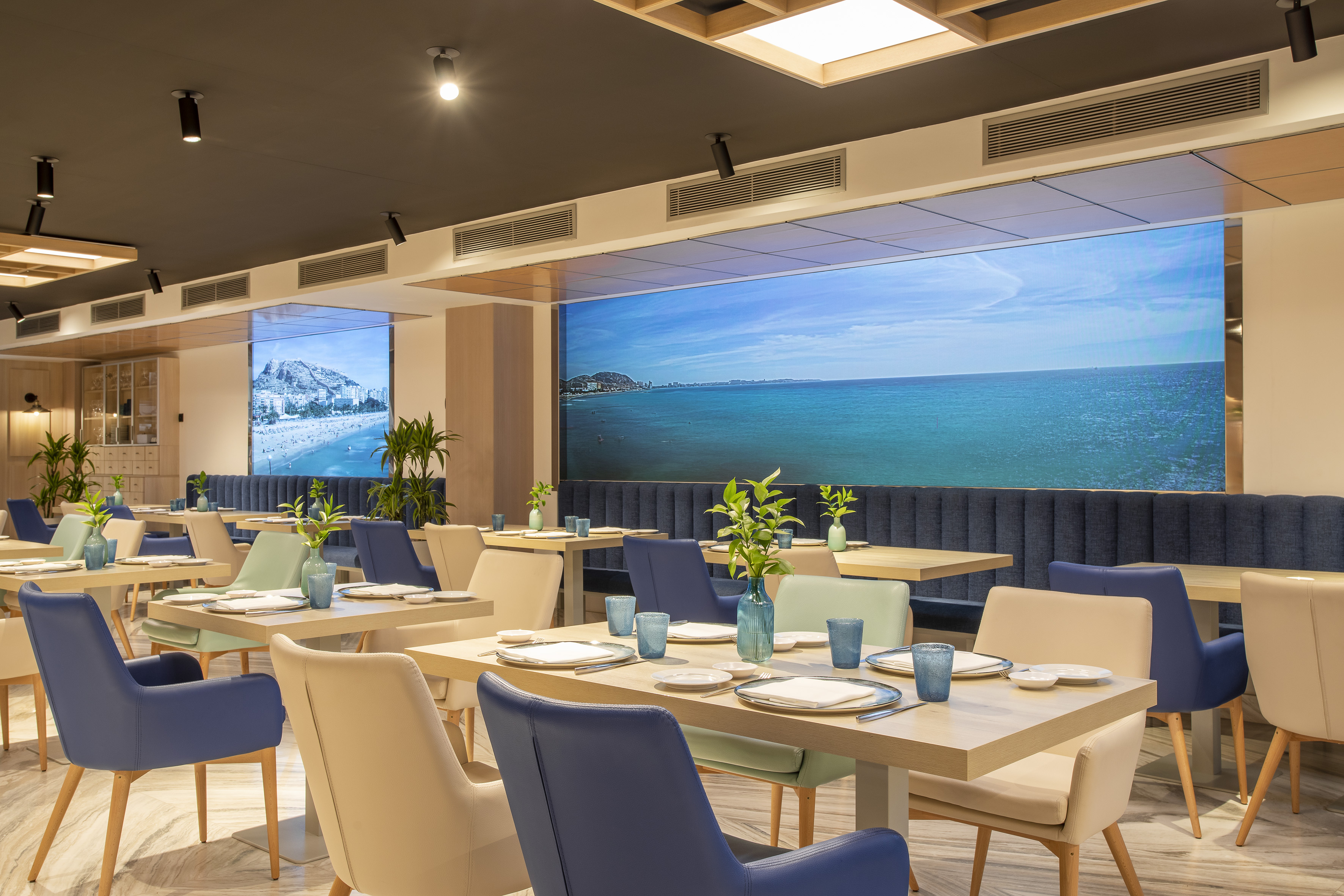 Modern restaurant with set tables, blue chairs, and large ocean view displays.