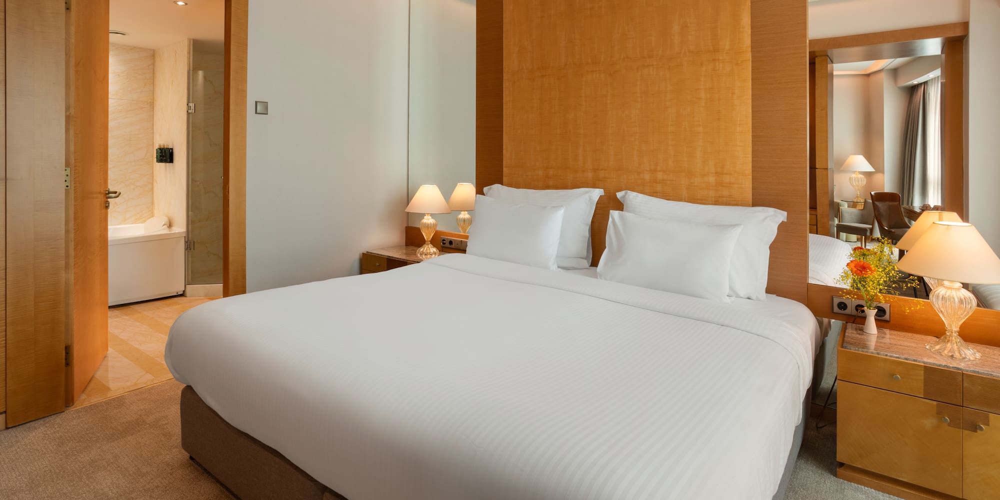 Modern hotel bedroom with king bed, wooden headboard, lamps, and bath.