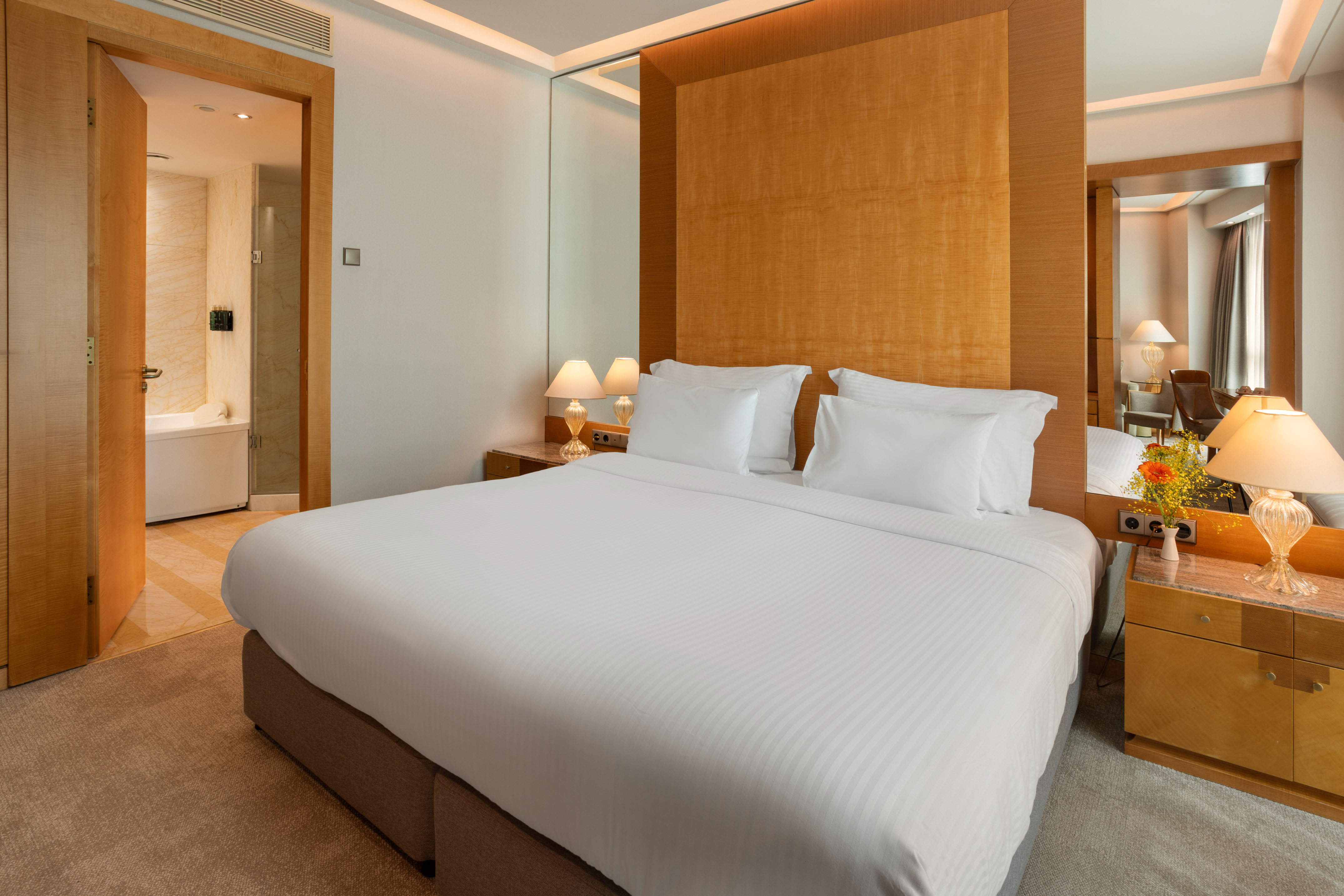 Modern hotel bedroom with king bed, wooden headboard, lamps, and bath.