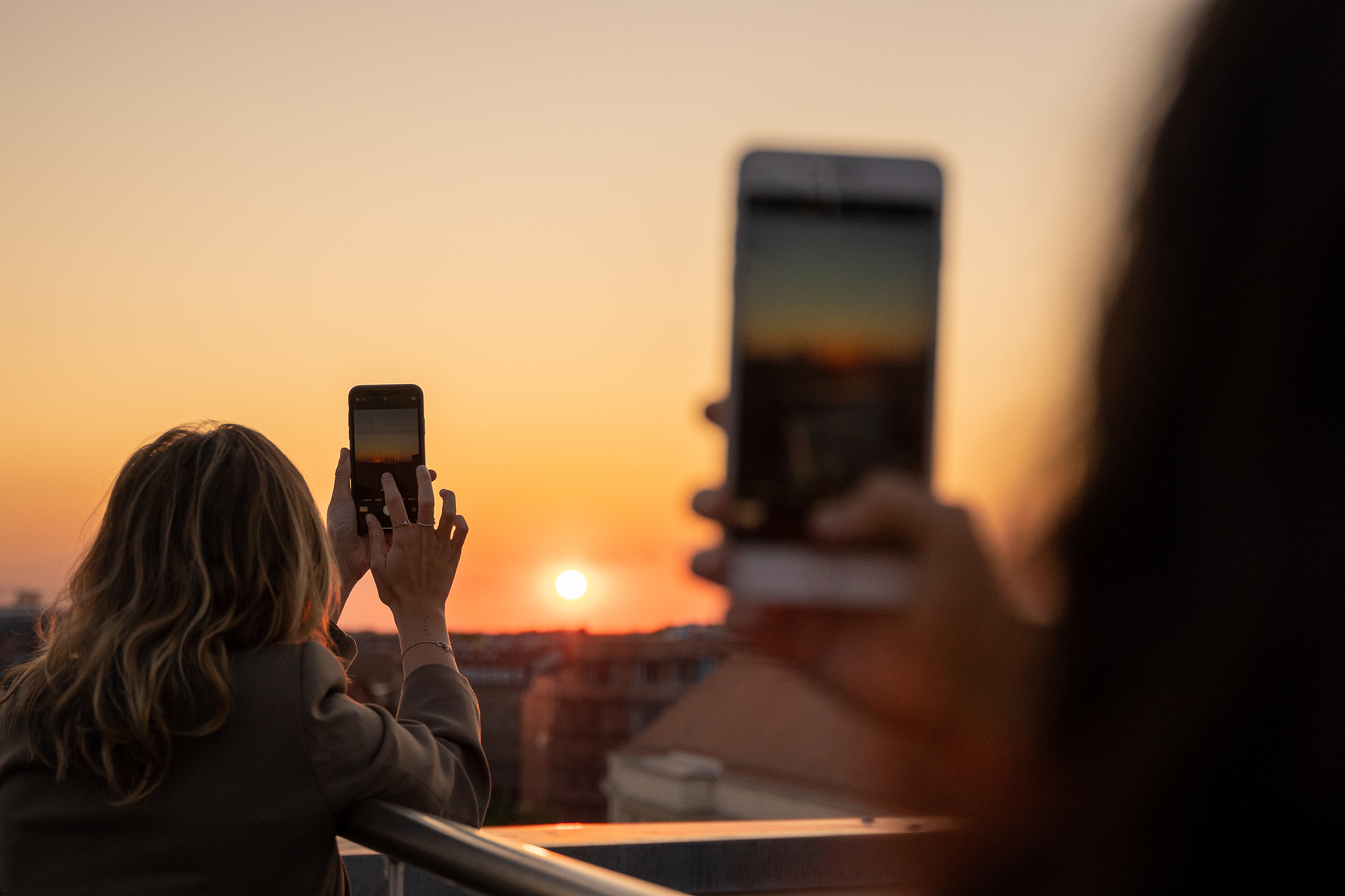 Woman photographing golden hour sunset over city with smartphone.