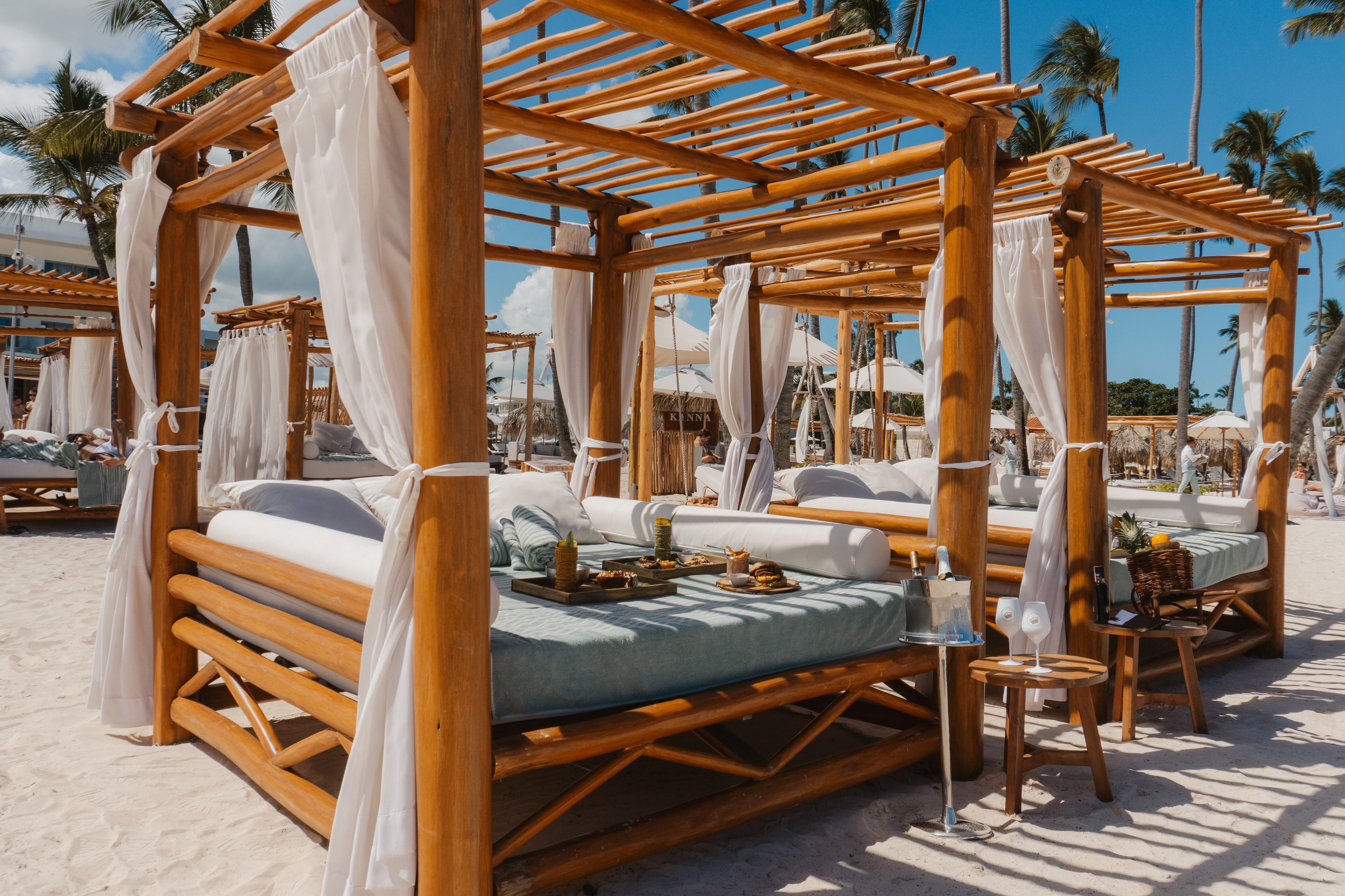 Luxury resort beach cabanas on sand with food, drinks, and palm trees.