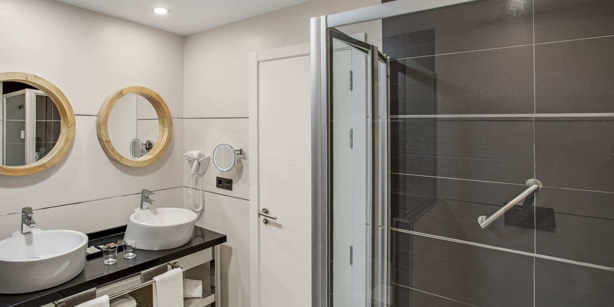Hotel bathroom with double vanity, wooden mirrors, and dark tiled shower.