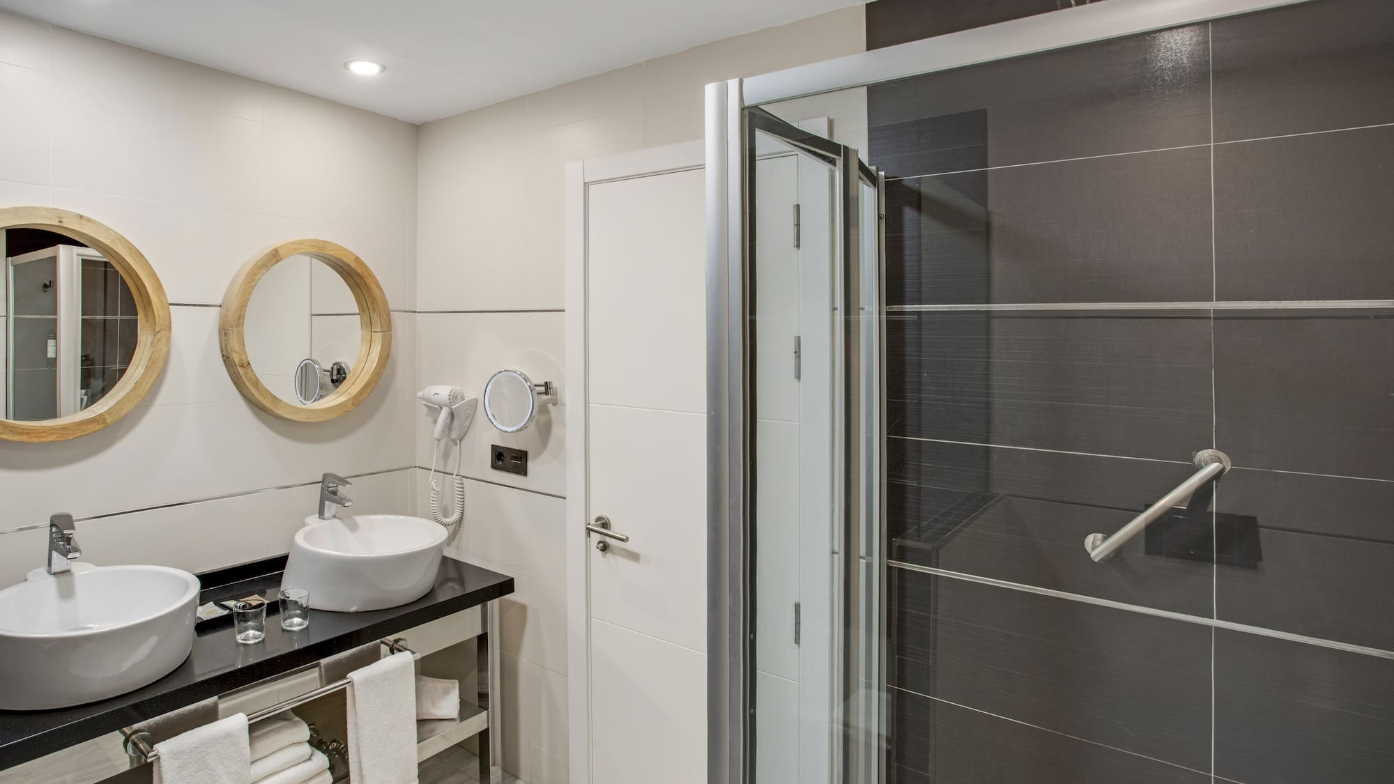 Hotel bathroom with double vanity, wooden mirrors, and dark tiled shower.