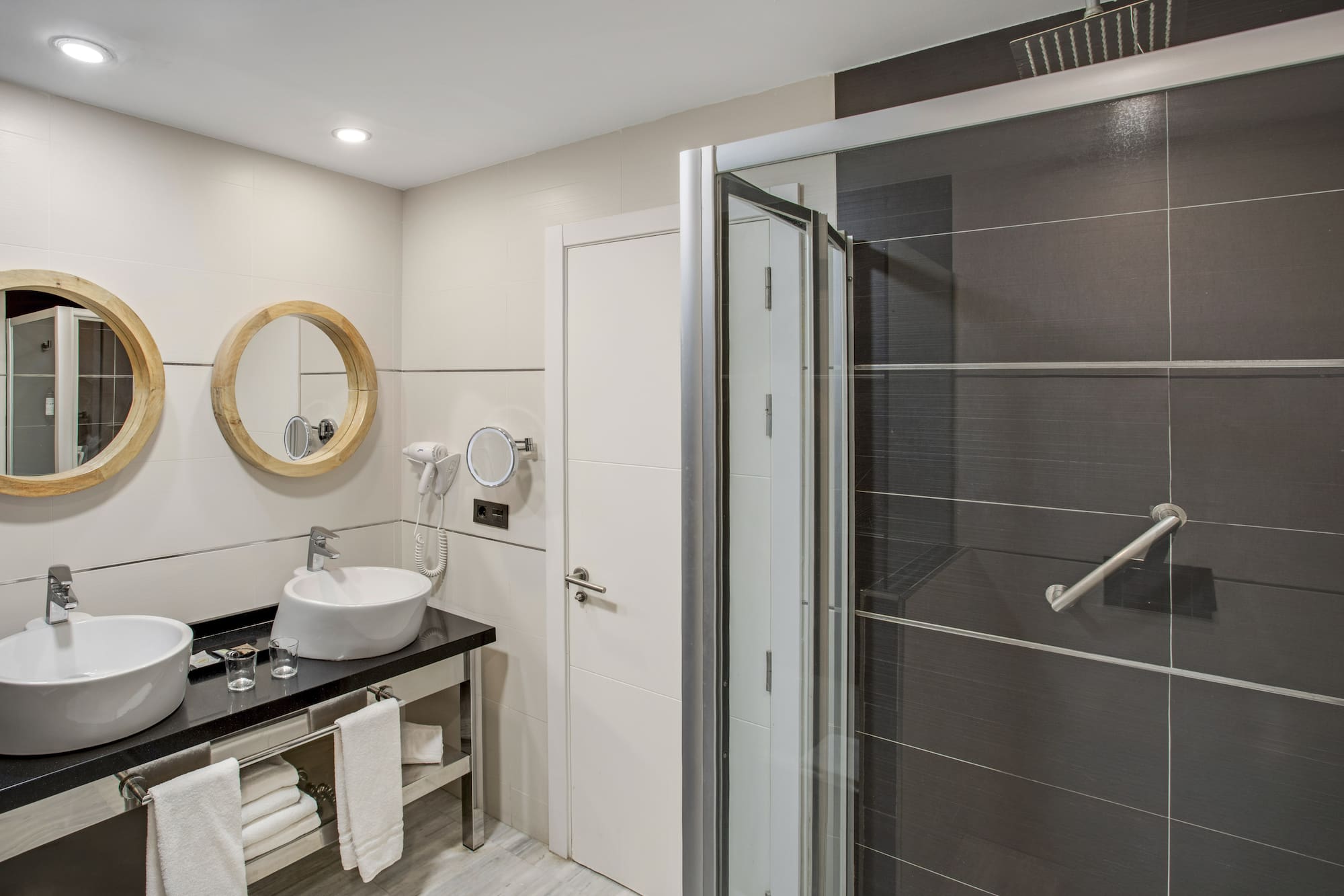Hotel bathroom with double vanity, wooden mirrors, and dark tiled shower.