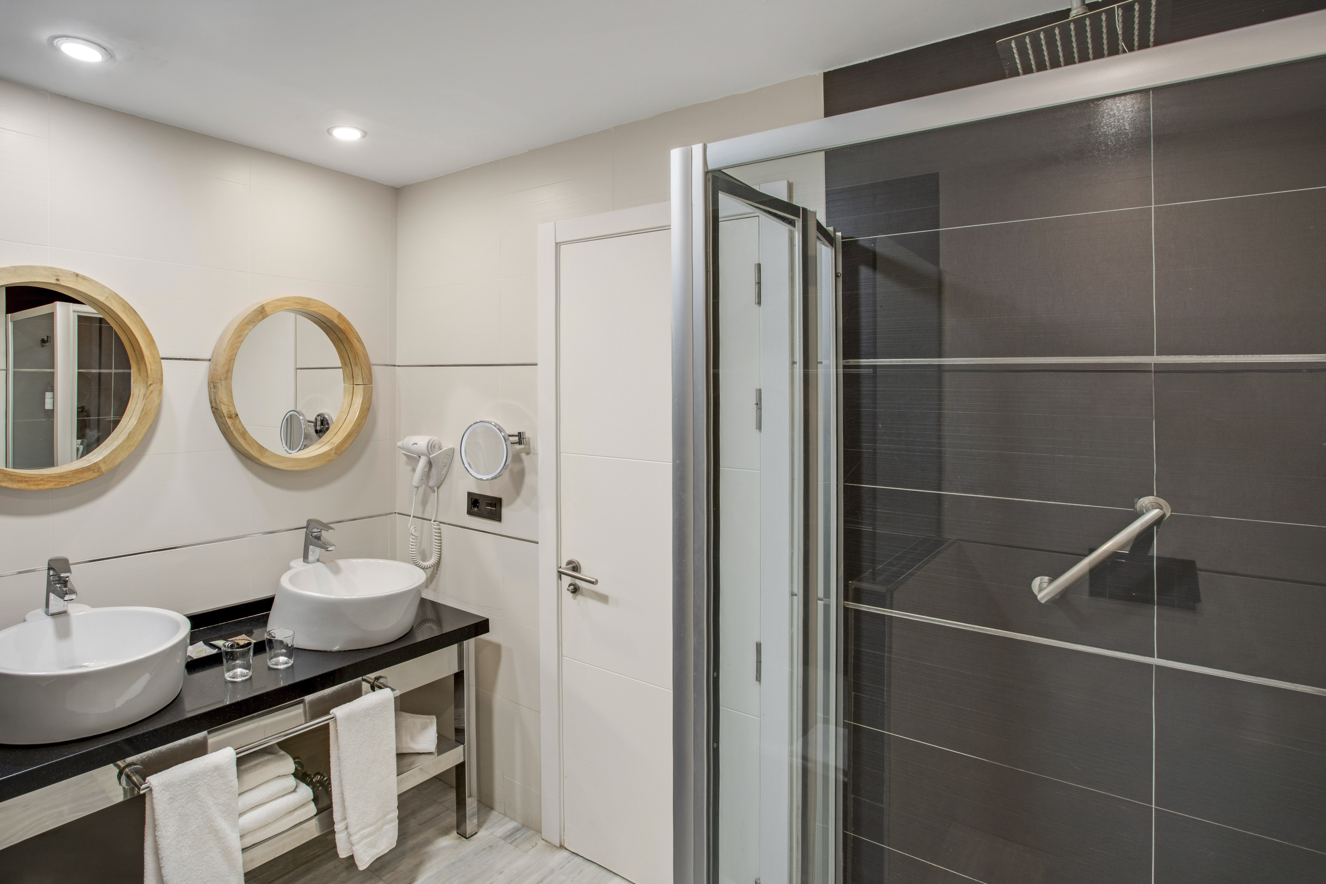 Hotel bathroom with double vanity, wooden mirrors, and dark tiled shower.