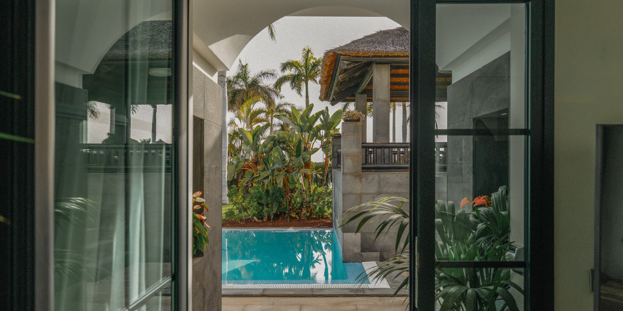 Tropical pool view from open doors, lush palms, and thatched roof villa.