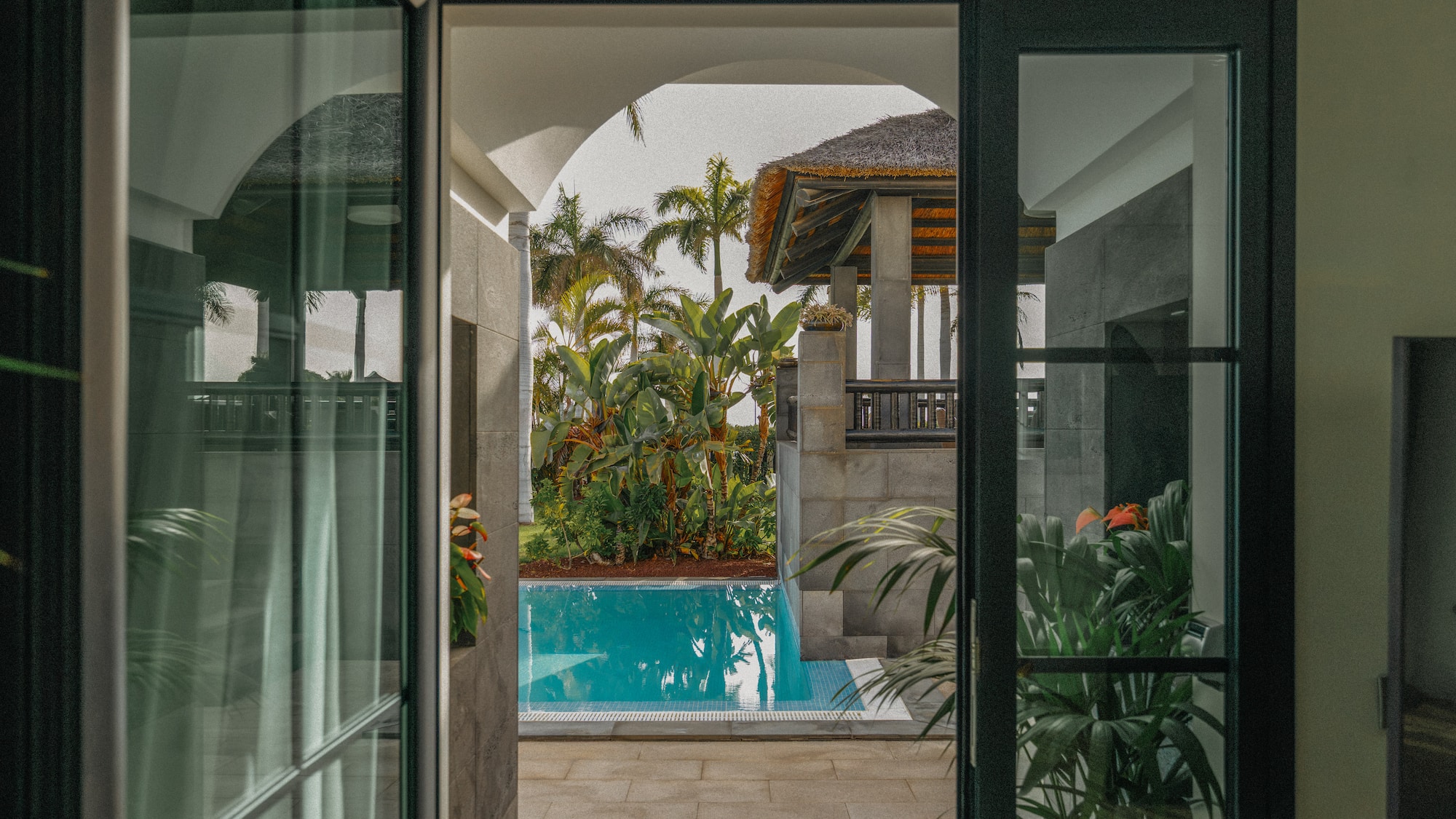 Tropical pool view from open doors, lush palms, and thatched roof villa.