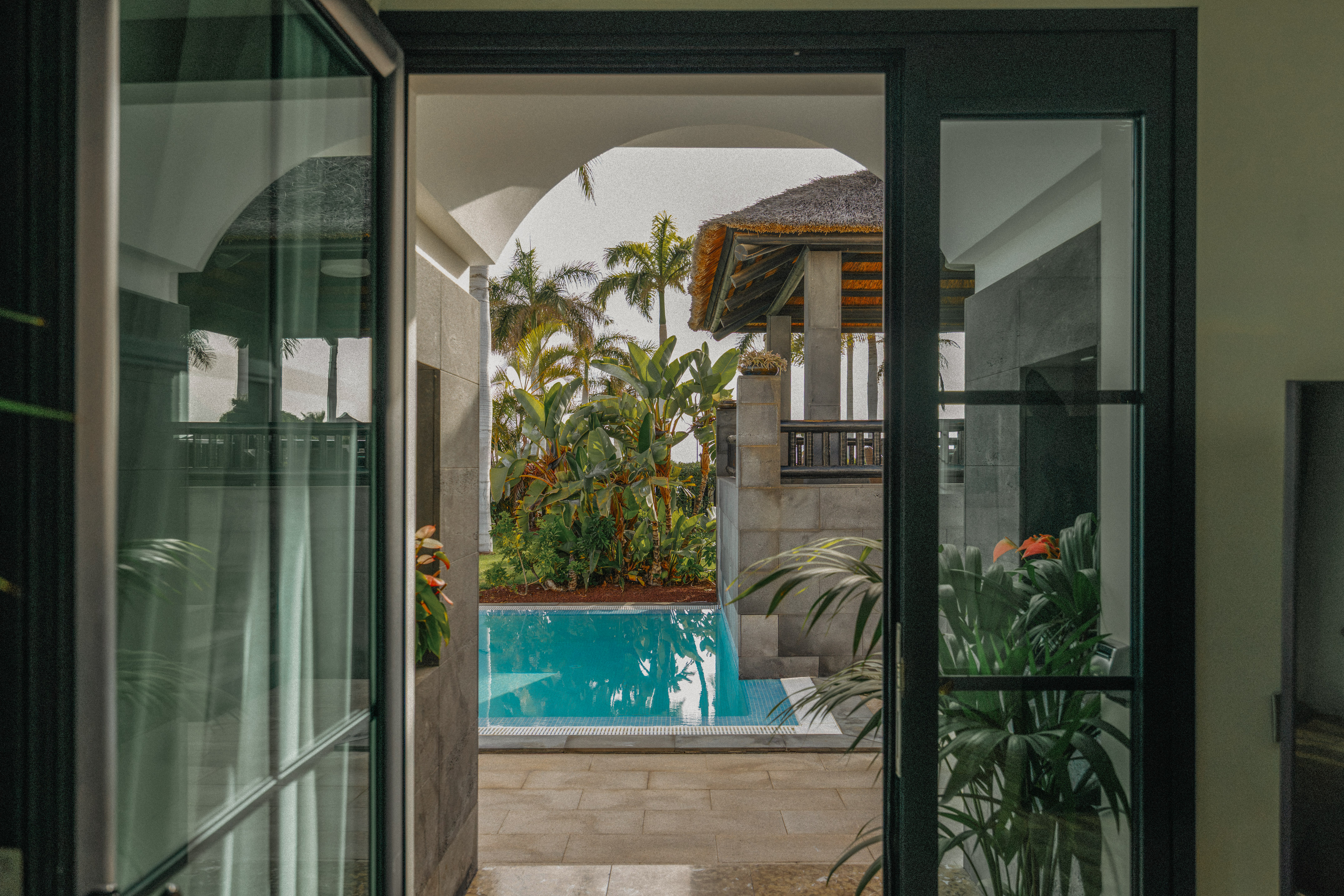 Tropical pool view from open doors, lush palms, and thatched roof villa.