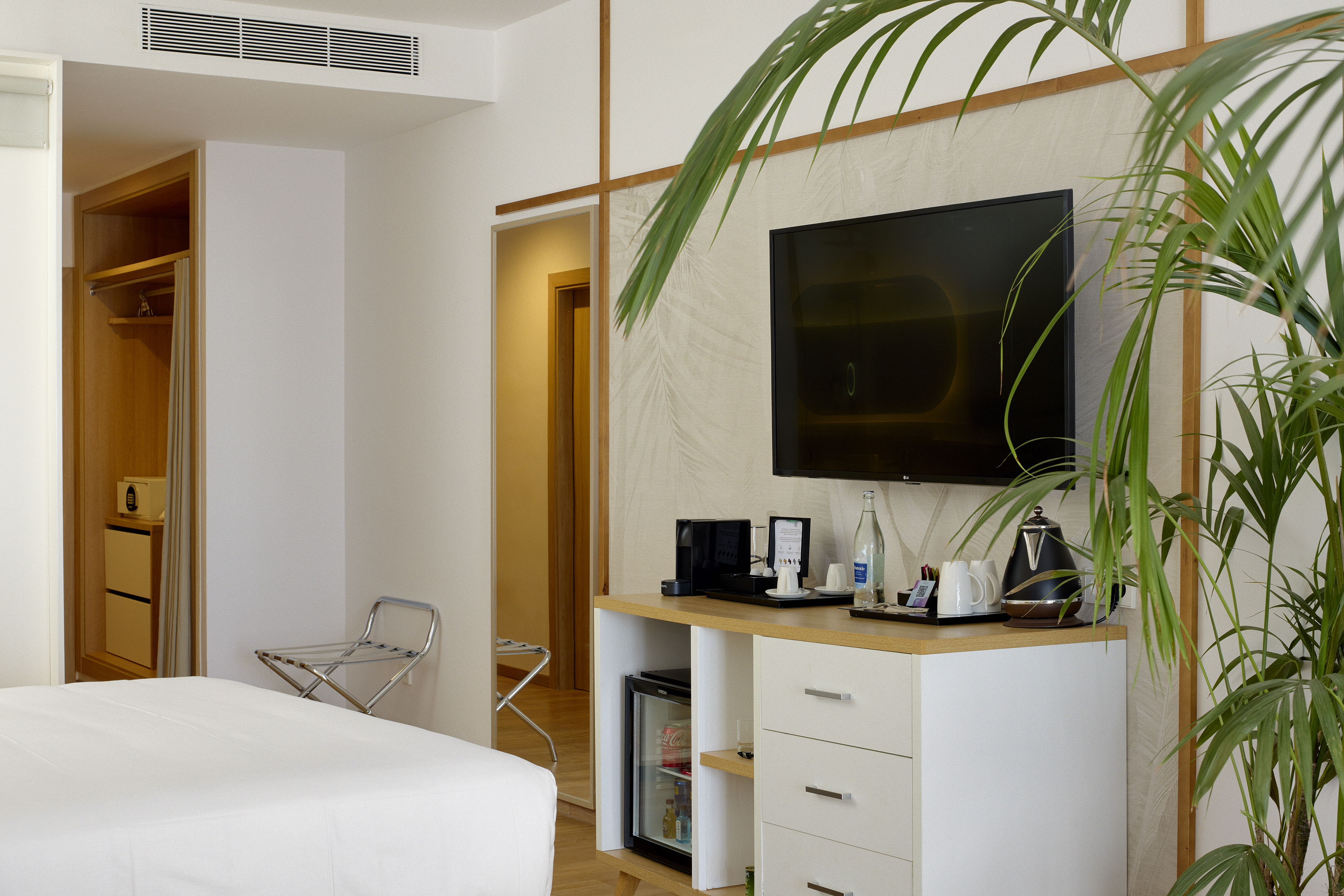 Modern hotel room: bed, TV, minibar, coffee station, and green plant.