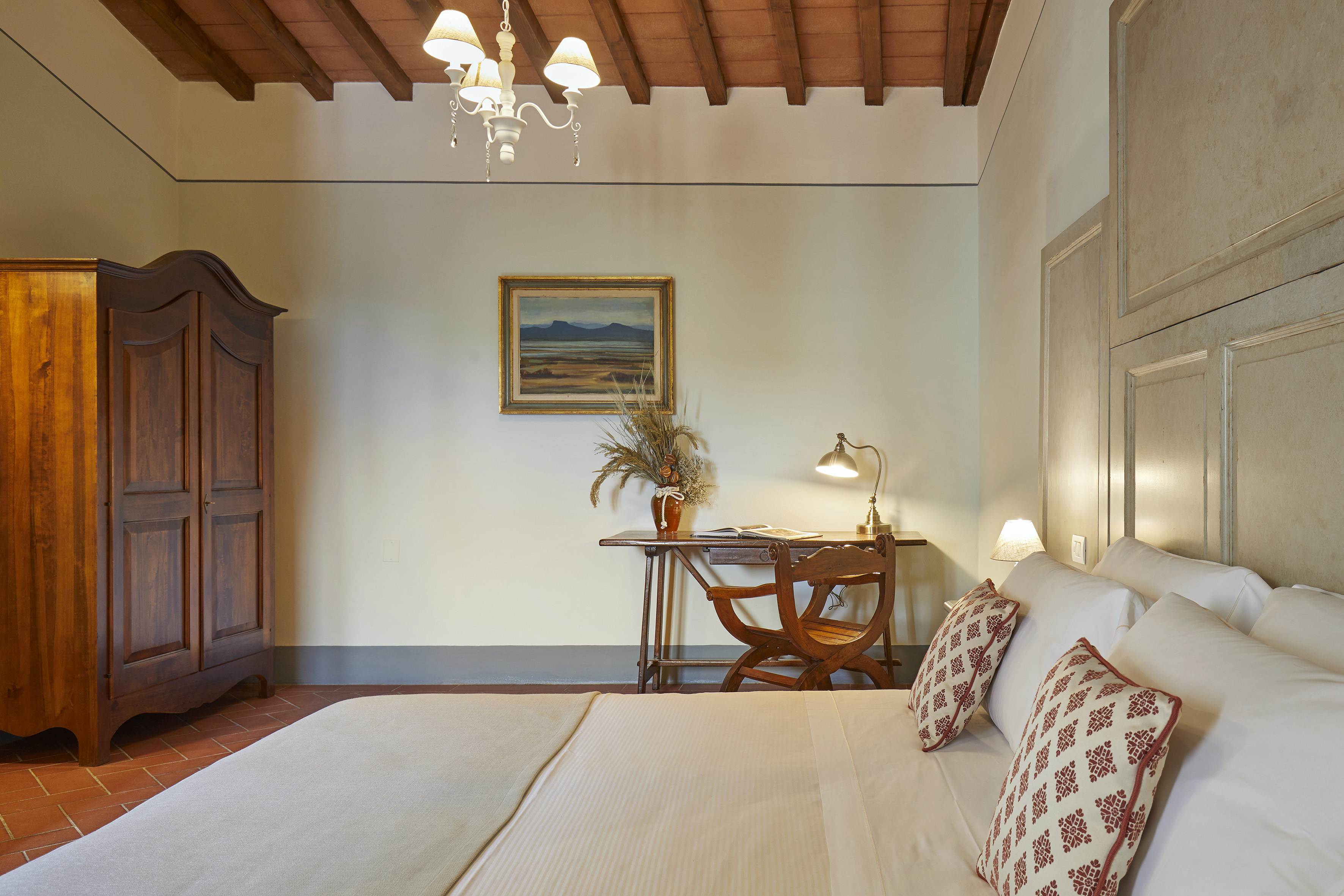 Rustic Tuscan bedroom with large bed, wooden wardrobe, desk, and chandelier.