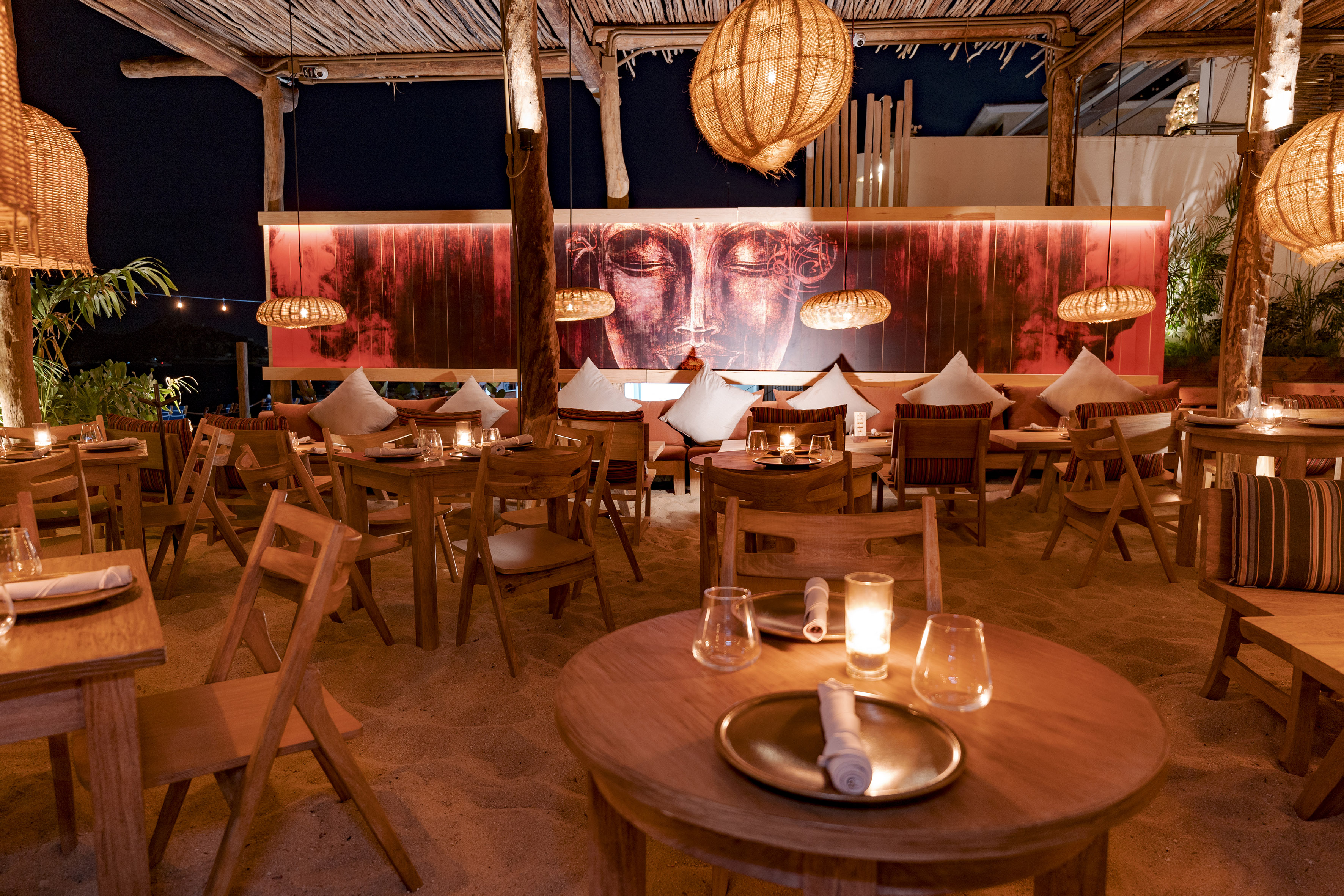 Tropical outdoor restaurant: sandy floor, Buddha art, woven lights at night.