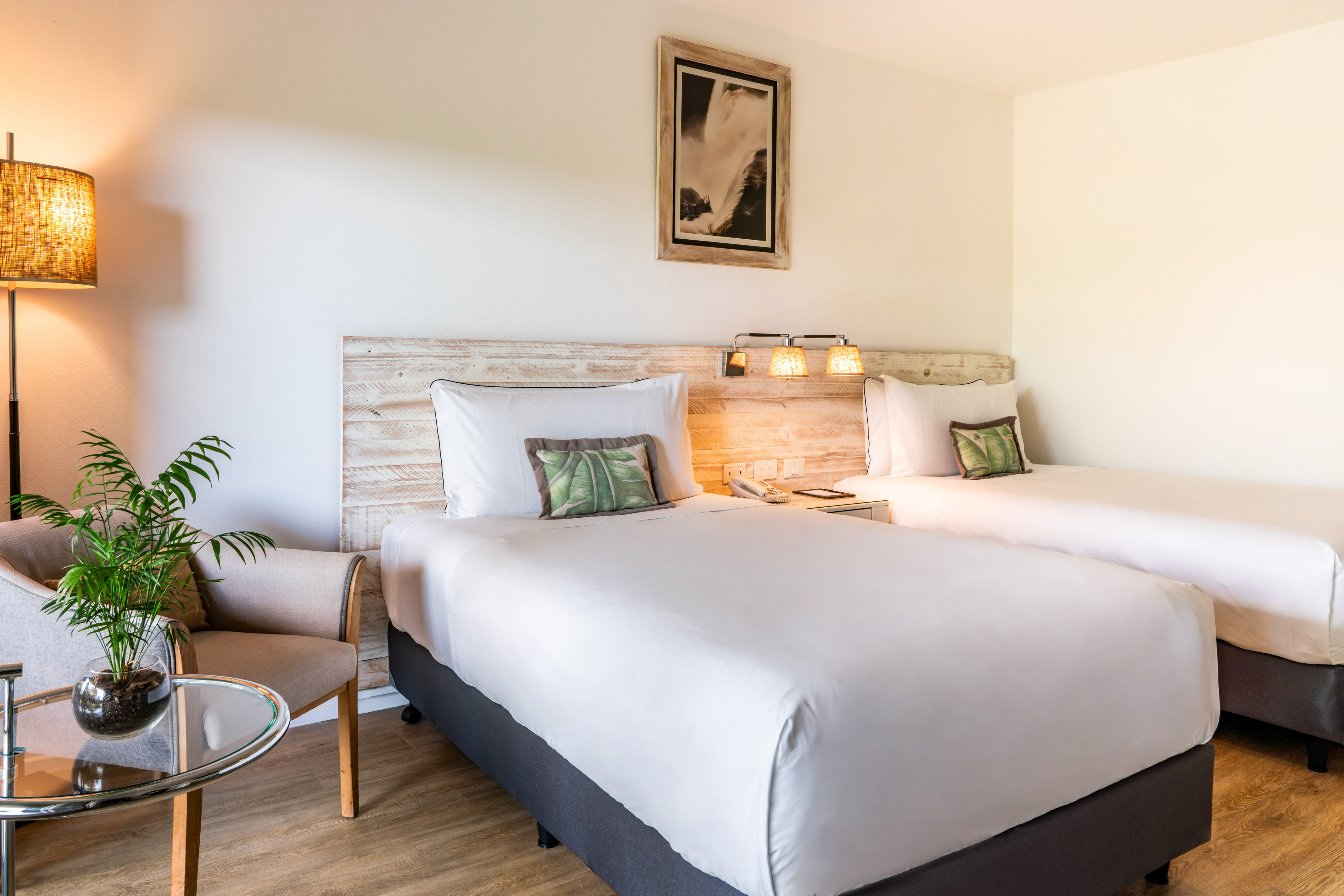 Cozy hotel room with two twin beds, wooden headboards, lamp, and tropical plant.