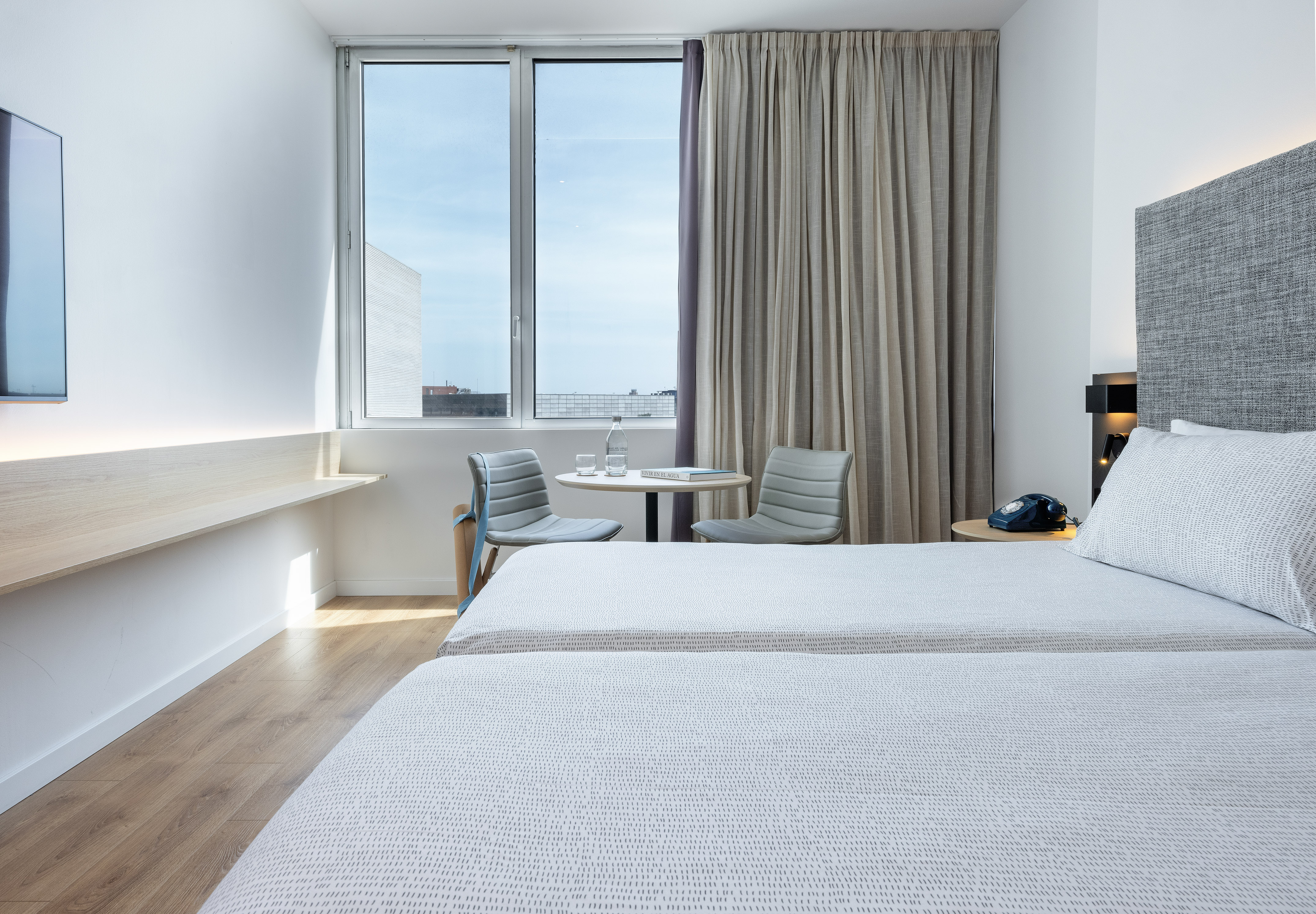 Modern hotel room with twin beds, bright window, wooden floor, and seating.
