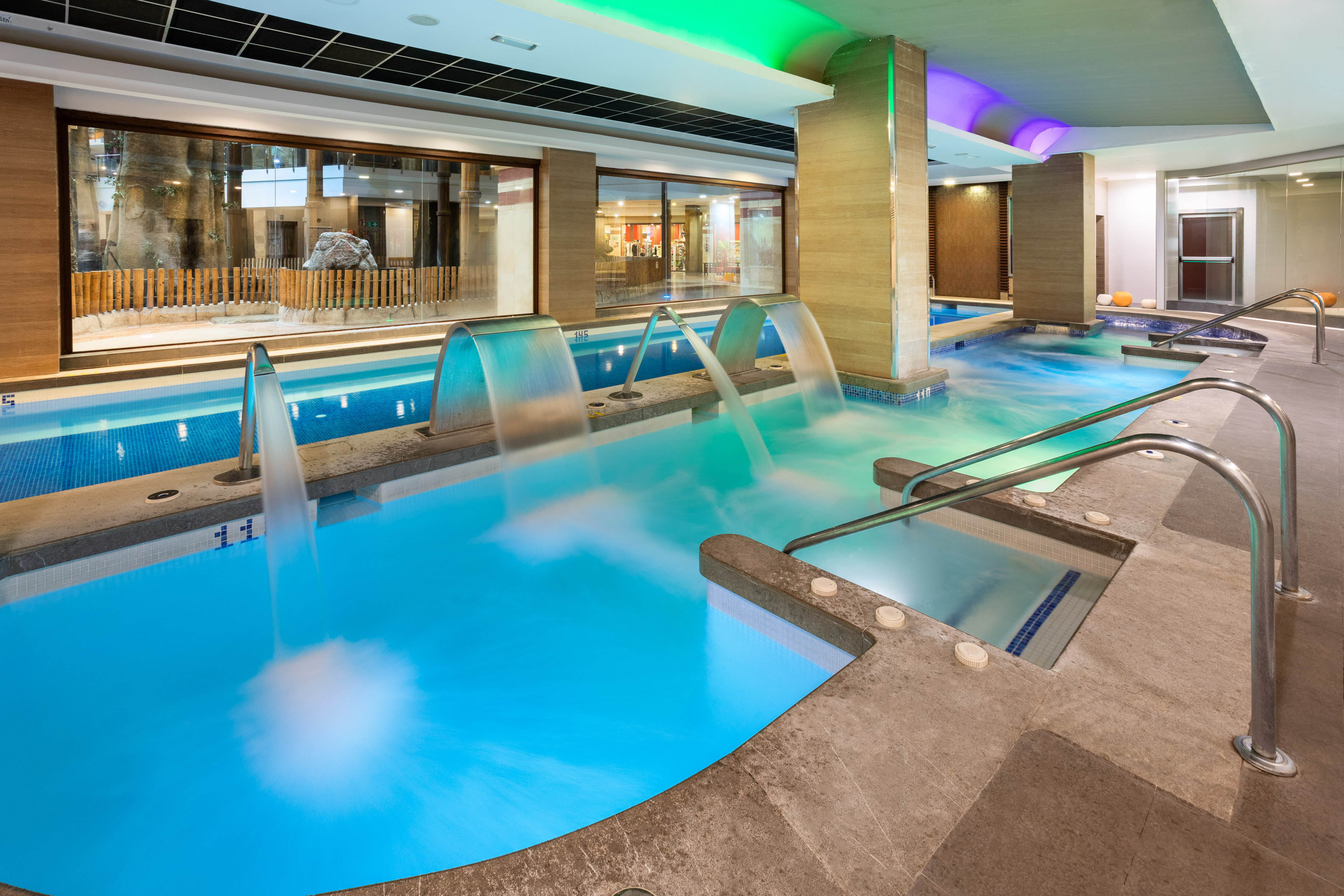 Luxury indoor spa pool with hydrotherapy jets, waterfalls, and colorful lighting.
