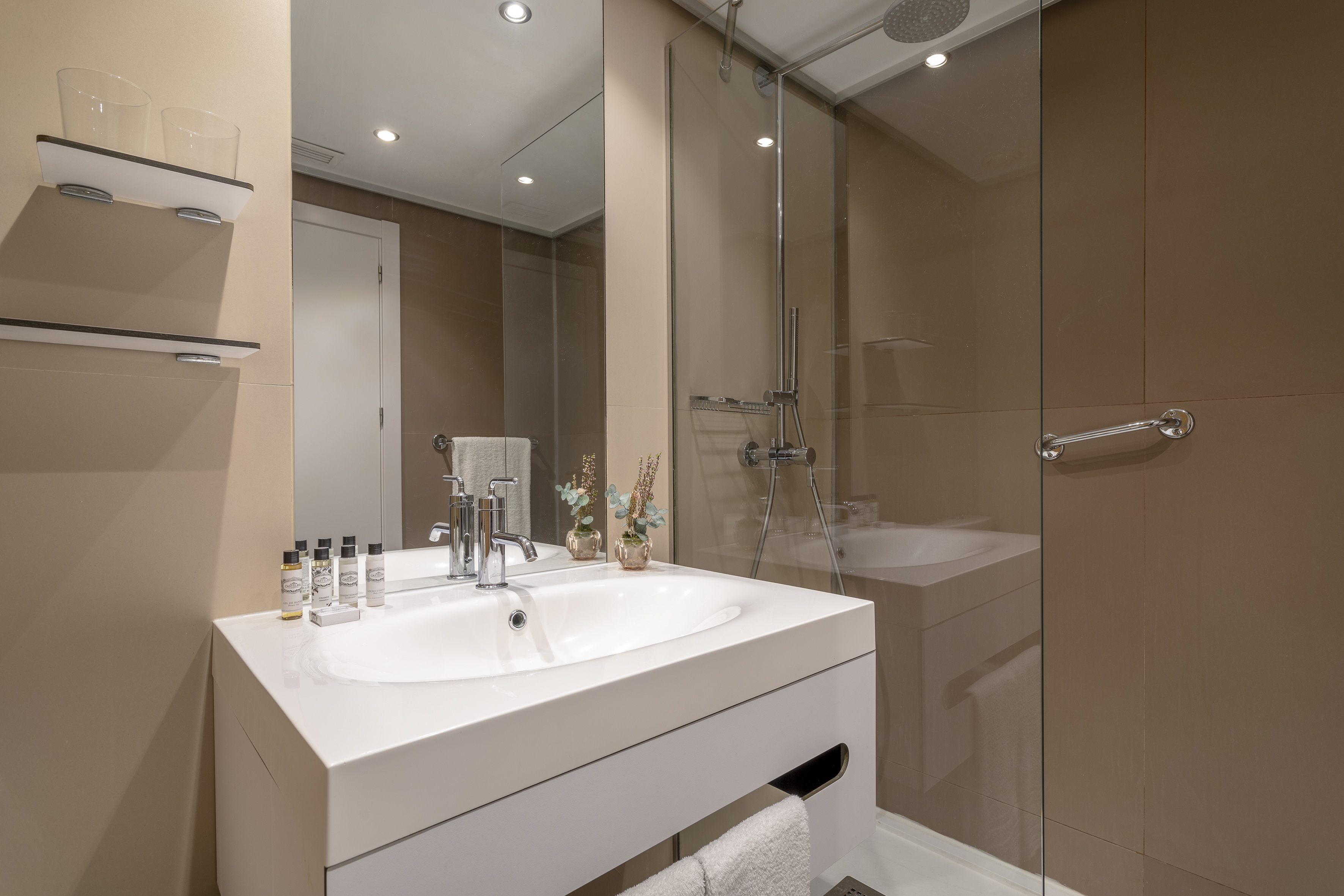 Modern hotel bathroom with white sink, mirror, glass shower, and beige tiled walls.