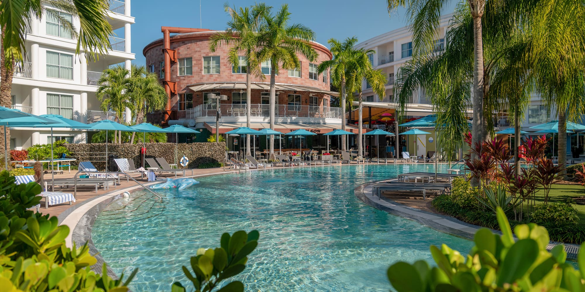 Sunny resort pool, palm trees, blue umbrellas, and hotel buildings.