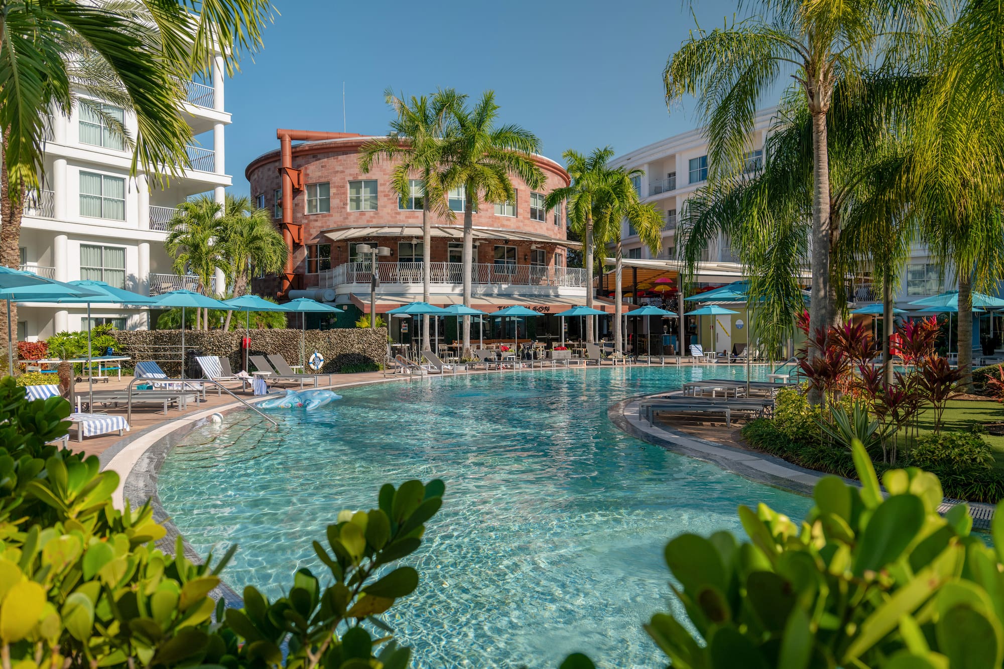 Tropical resort pool with palm trees, blue umbrellas, and hotel buildings.