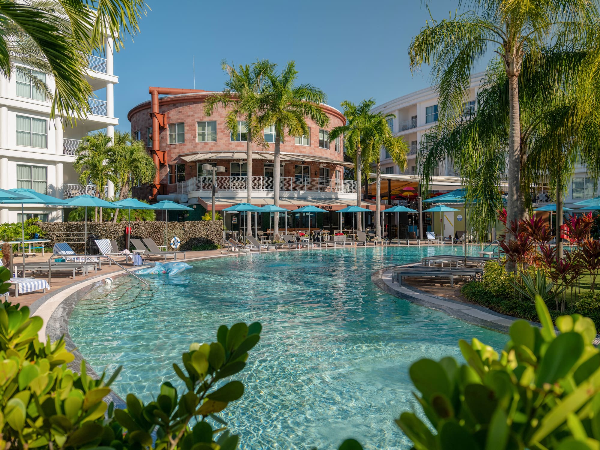 Sunny resort pool, palm trees, blue umbrellas, and hotel buildings.