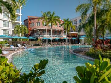 Tropical resort pool with palm trees, hotel buildings, and blue umbrellas.