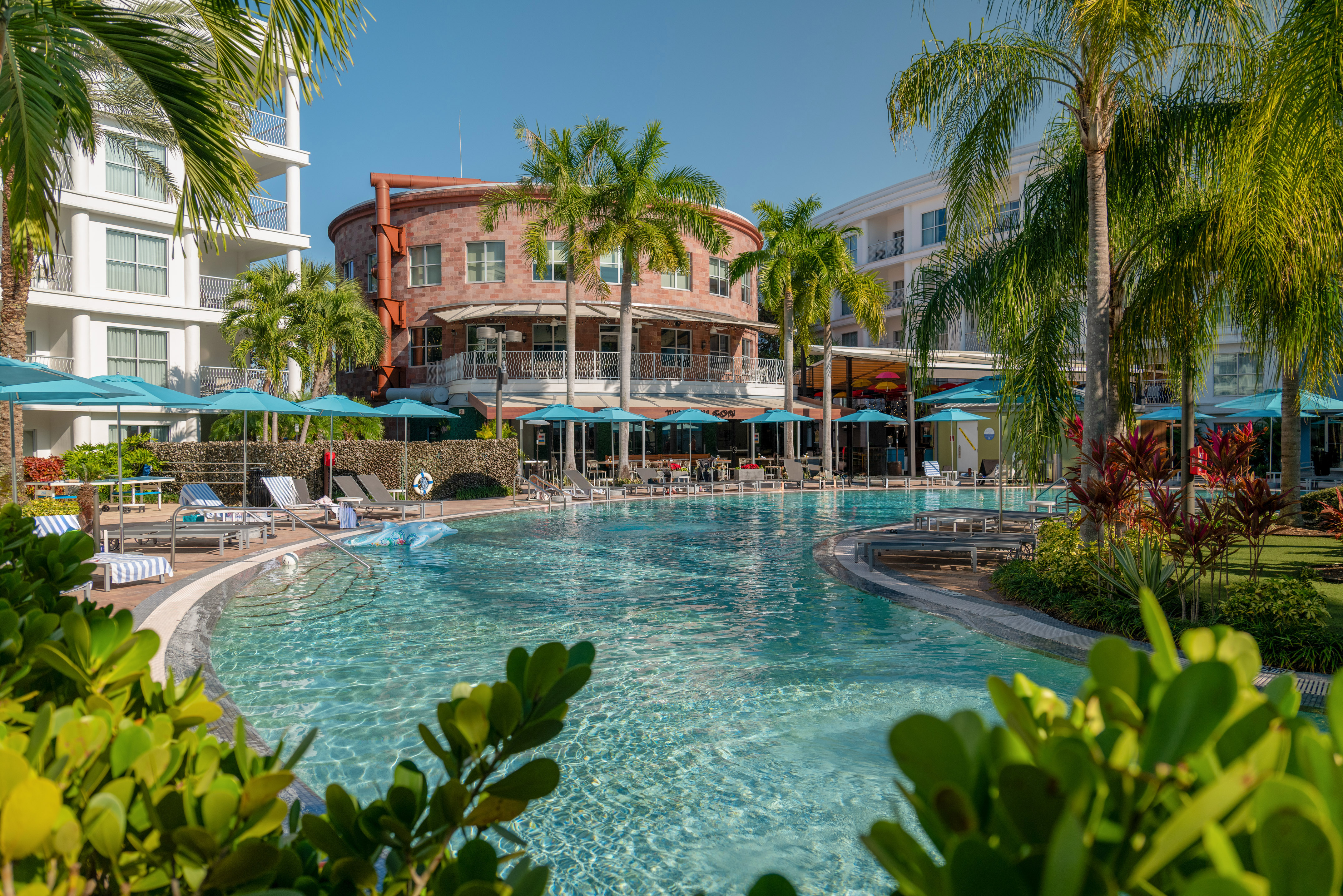 Tropical resort pool with palm trees, blue umbrellas, and hotel buildings.