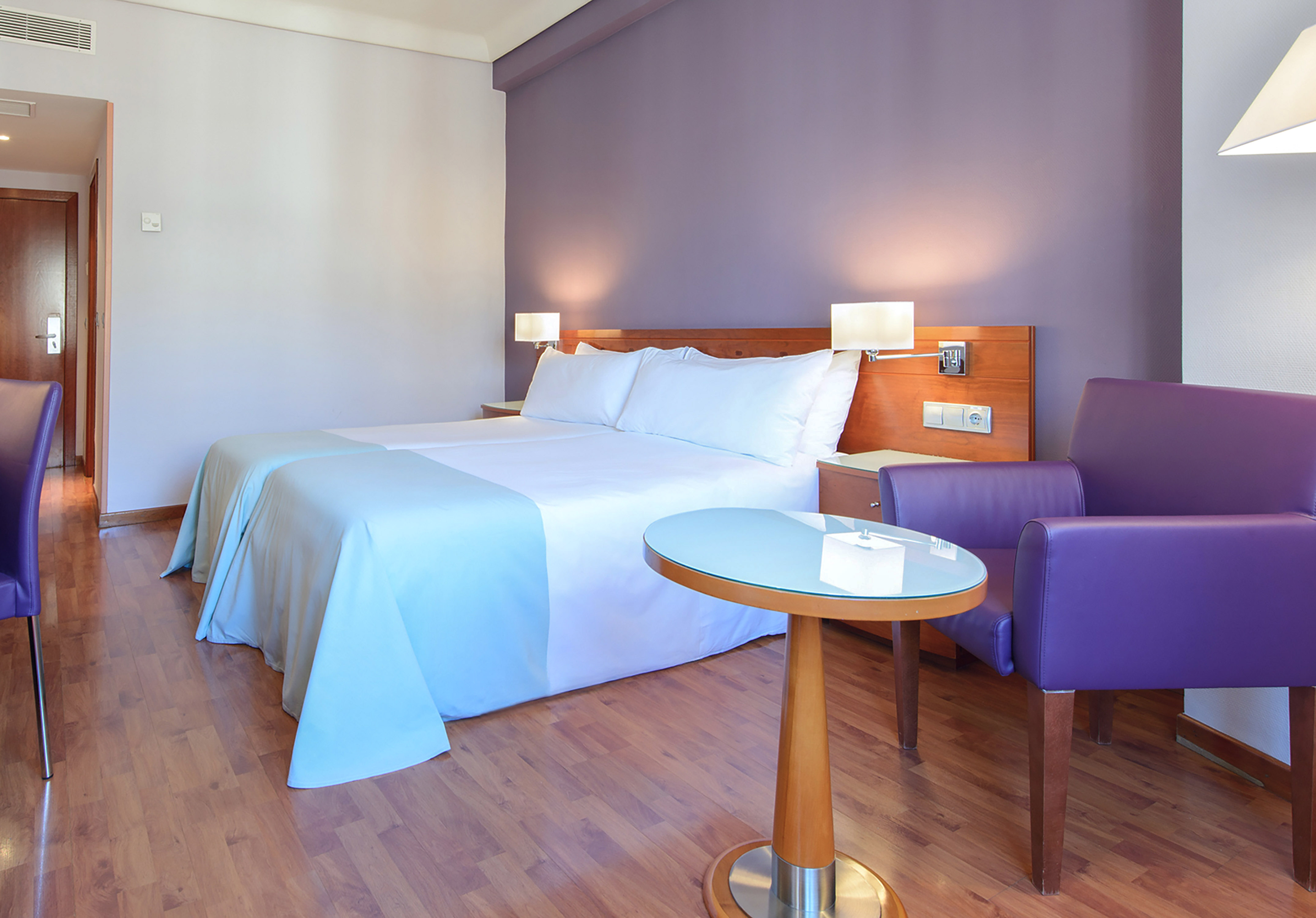 Hotel room with twin beds, purple accent wall, wood floor, armchair, and table.