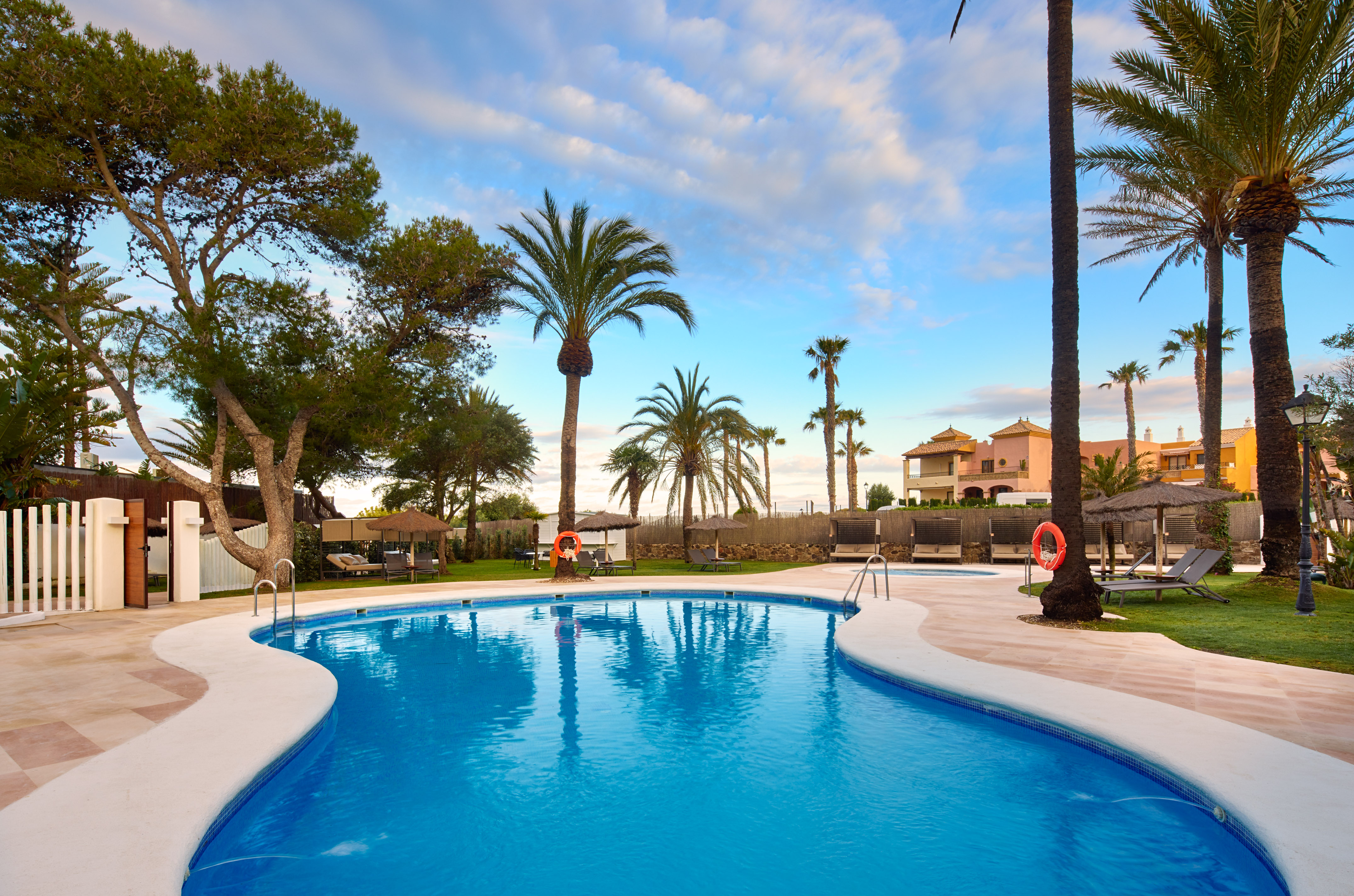 Luxury resort swimming pool with palm trees, lounge chairs, and hotel buildings.